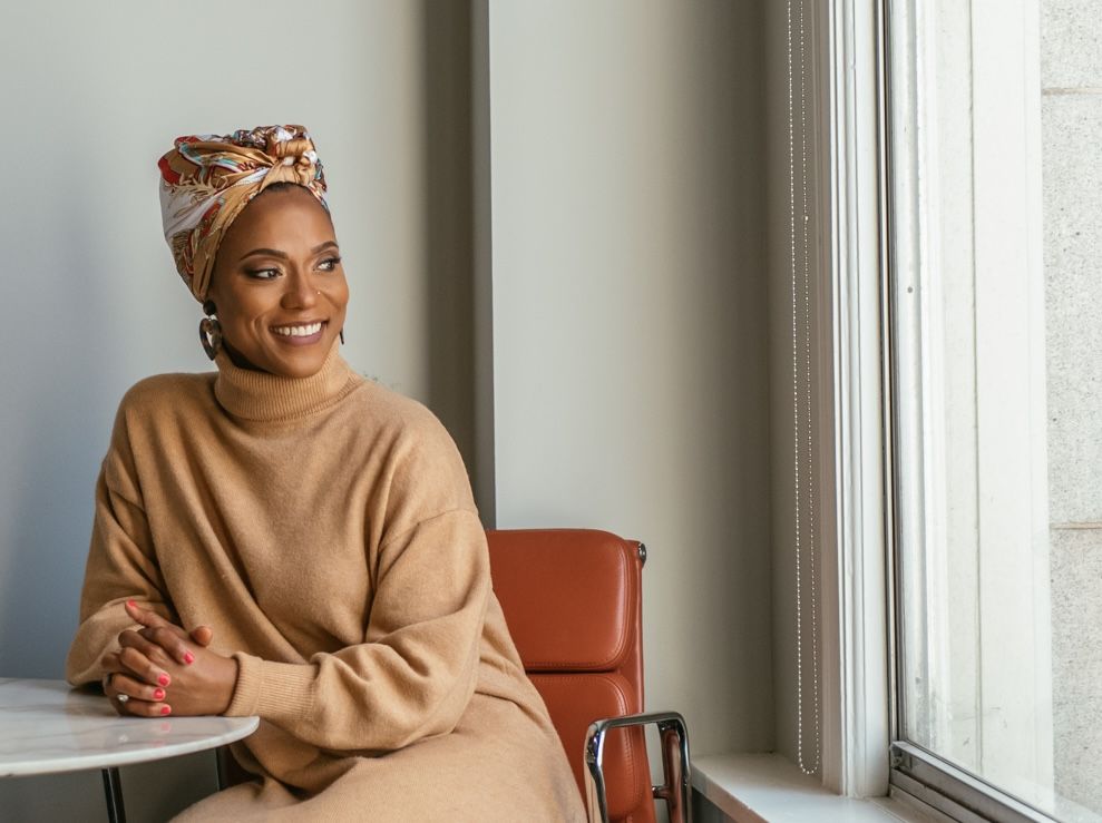 Smiling Black woman in beige sweater and colorful head wrap sits by window in modern interior.