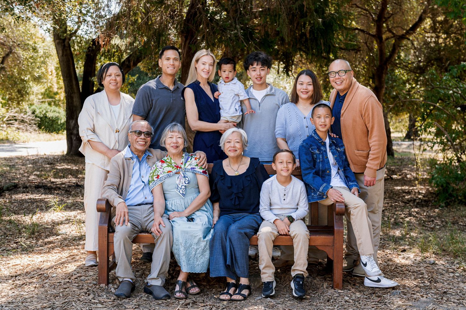 Multi-generational Asian family portrait outdoors, three rows of smiling relatives posing together among trees in warm natural light.