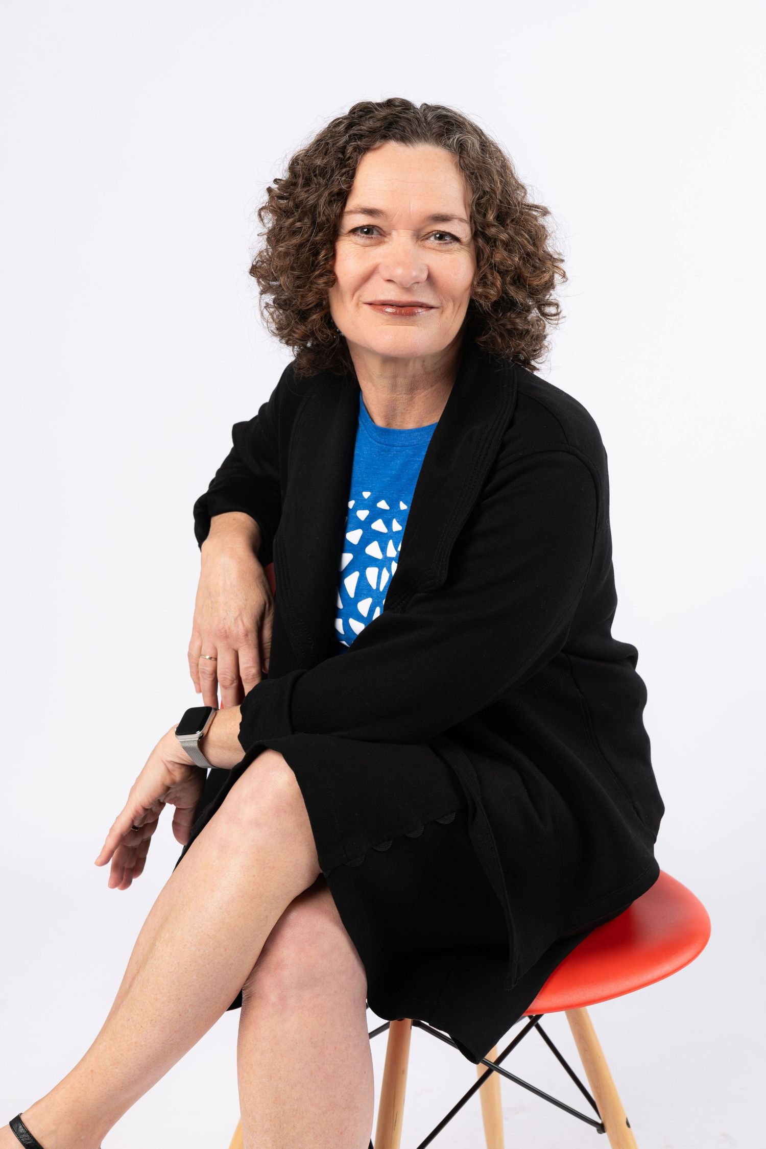 Professional woman with curly hair sitting on red chair, wearing black blazer and blue top, white background.
