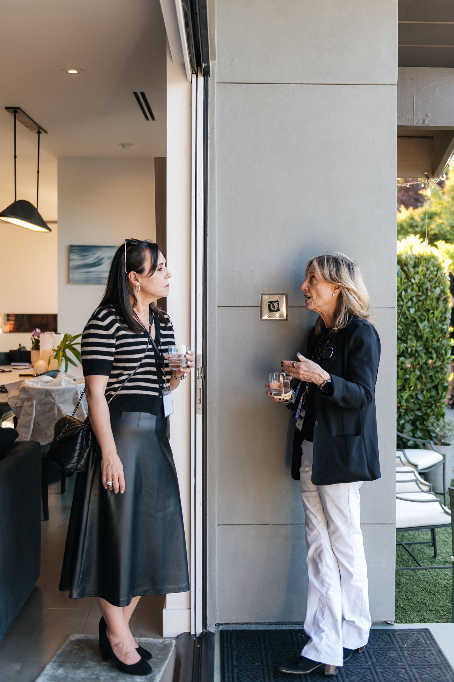 Two women chat by a modern home entrance, holding drinks, one in striped top and leather skirt, other in black blazer.