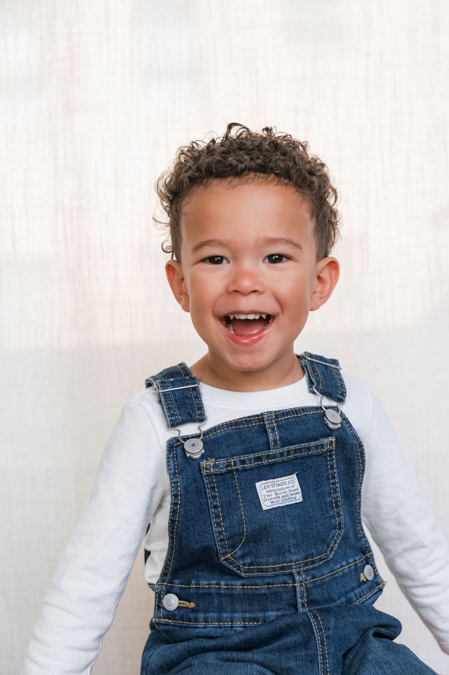 Smiling toddler with curly hair wearing denim overalls and white long-sleeve shirt against light background.