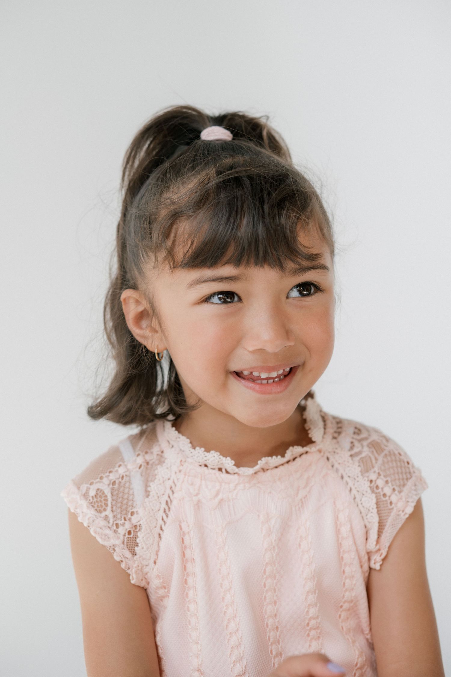 Smiling young girl with dark hair in ponytail wearing pink lace top against white background.