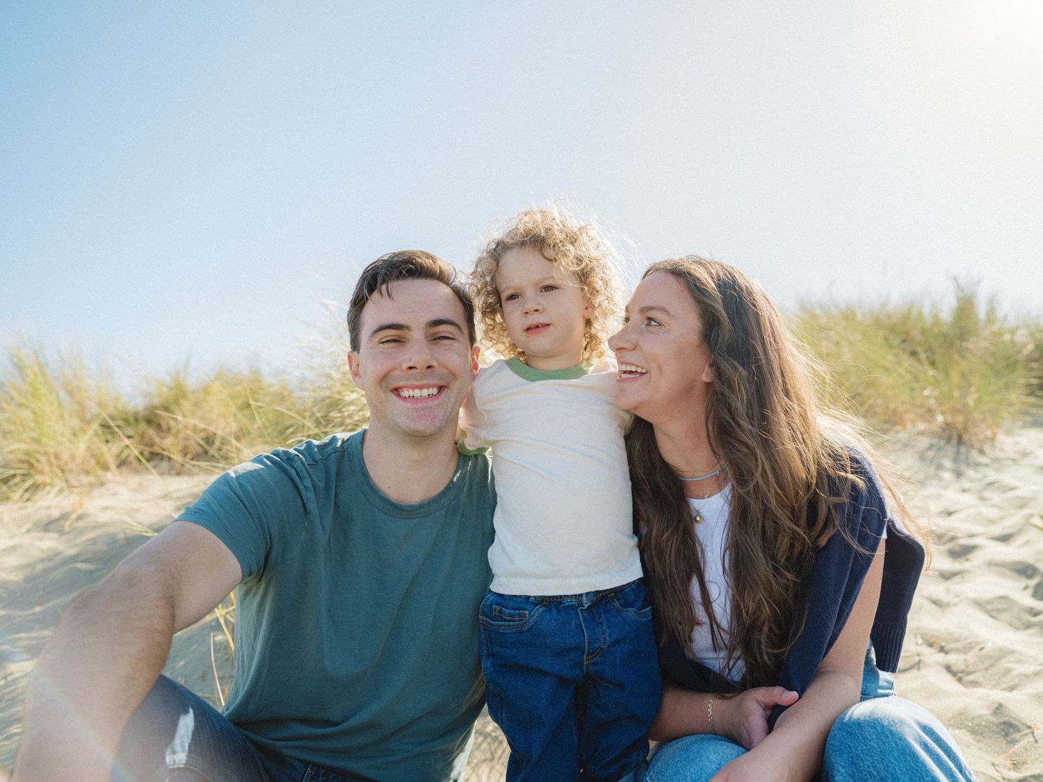 Happy family with young child smiling together on a sunny beach with sand dunes and blue sky.