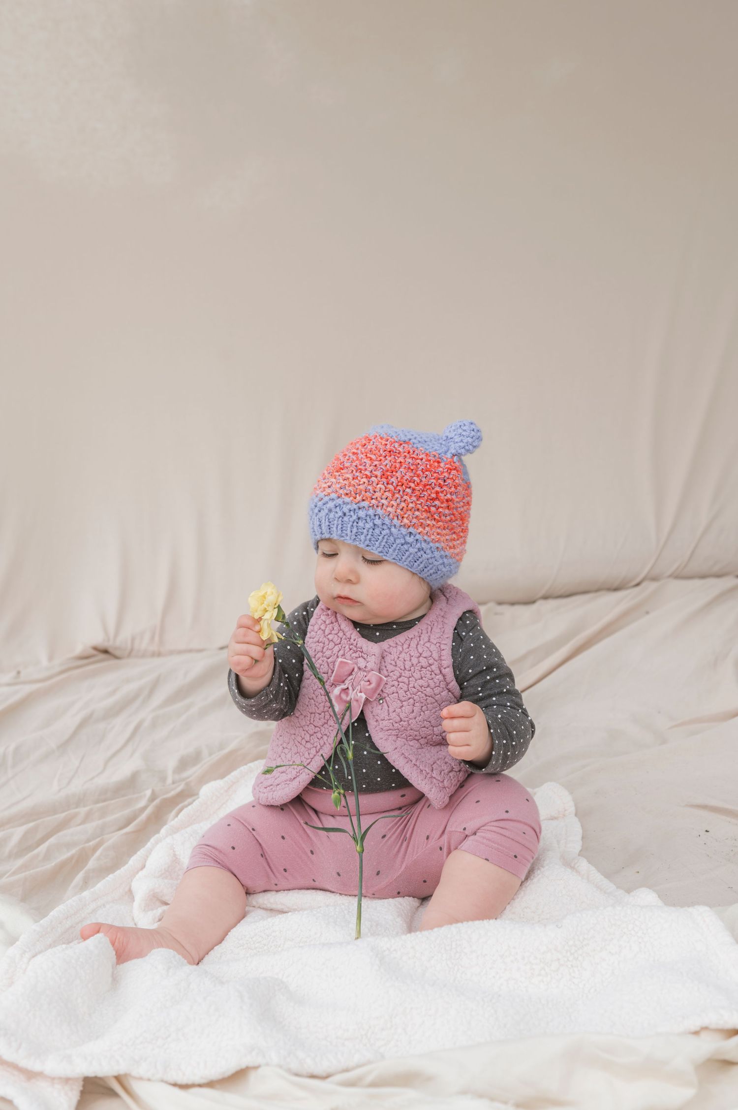 Baby wearing colorful knit hat and pink vest, holding a small yellow flower while sitting on a white blanket.