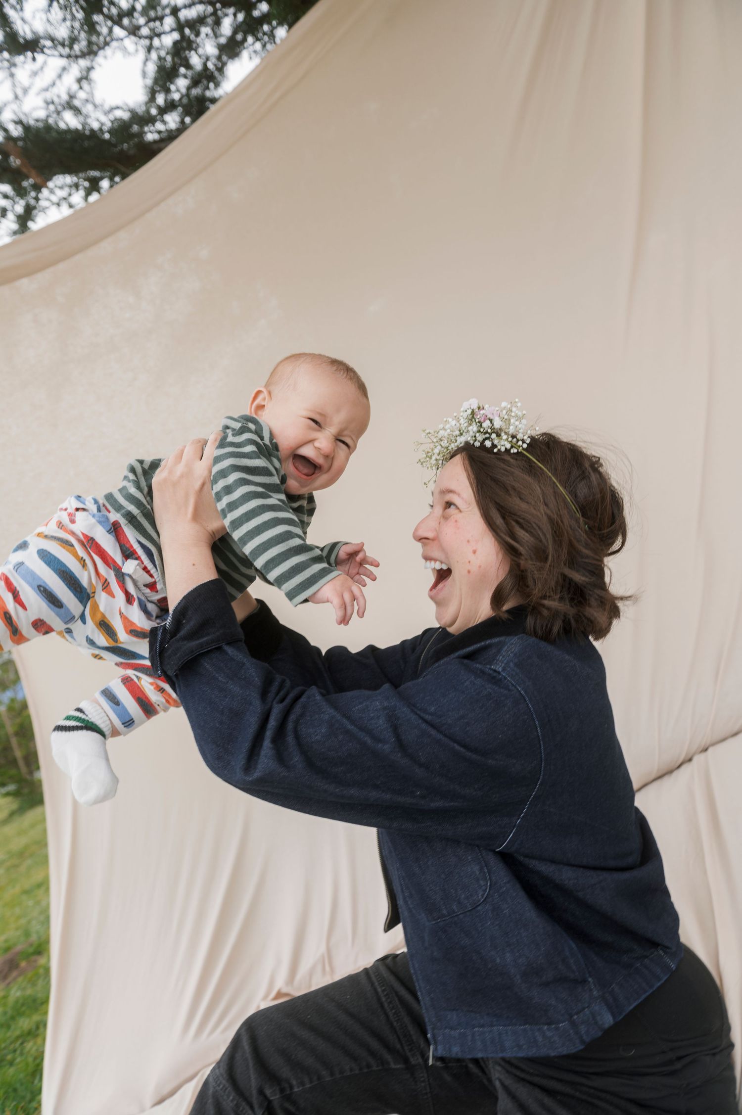 A joyful woman wearing a flower crown lifts a laughing baby outdoors near a white fabric backdrop.