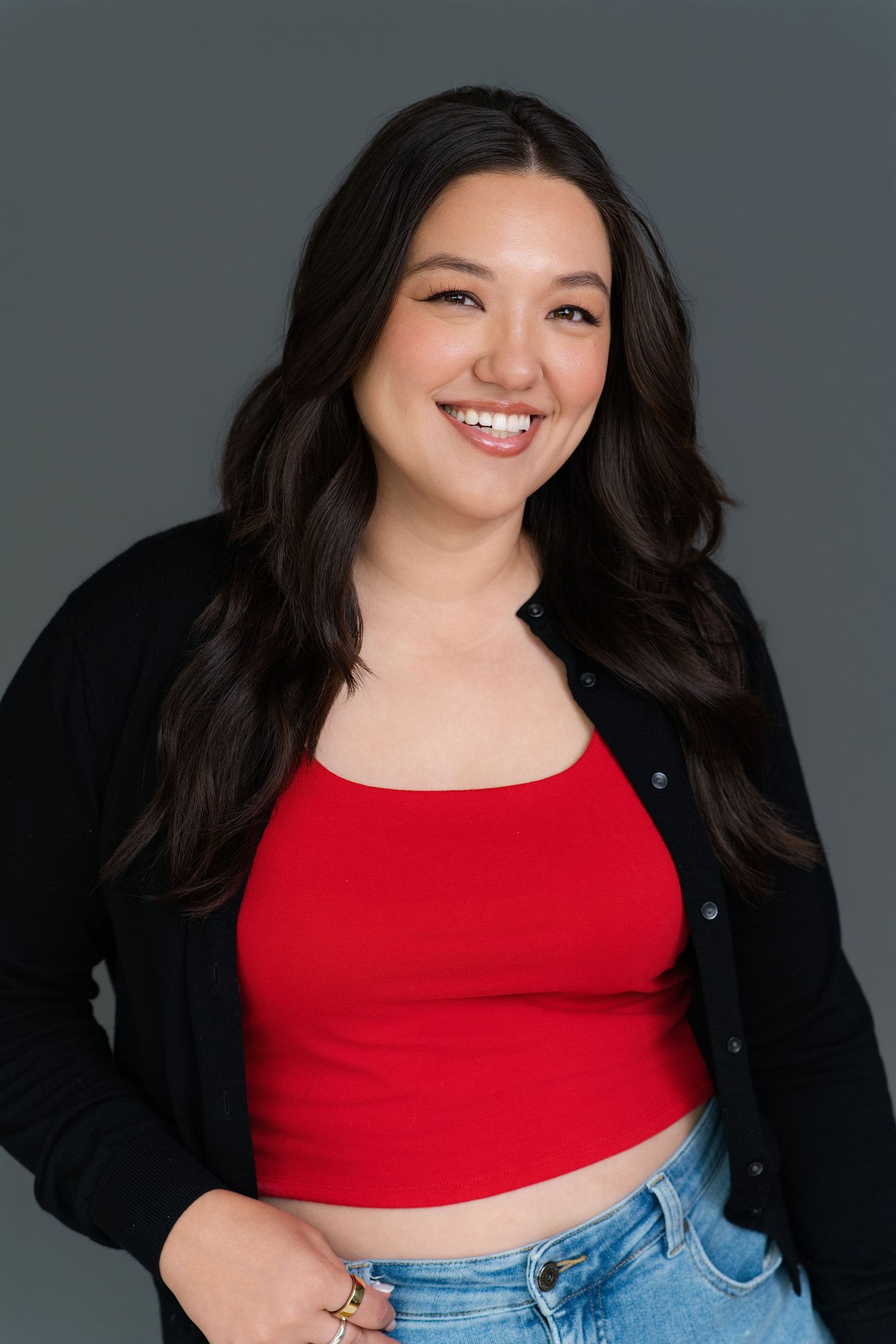 Smiling young woman with long dark hair wearing a red crop top, black cardigan, and jeans against a gray background.