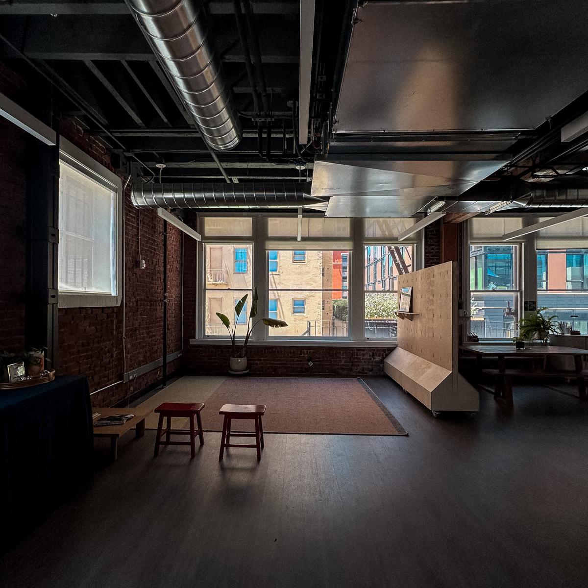 Industrial loft space with exposed brick walls, ductwork ceiling, large windows, wooden bench, and dark hardwood floors.