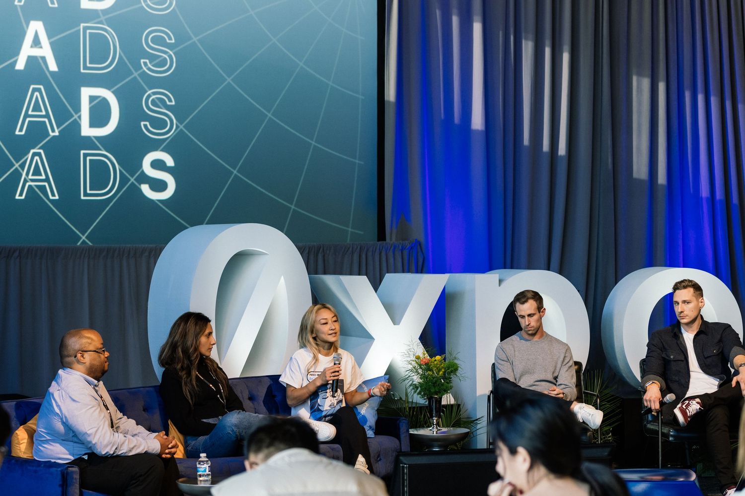 Panel discussion on stage at ADS EXPO conference with five speakers seated in blue chairs against branded backdrop.