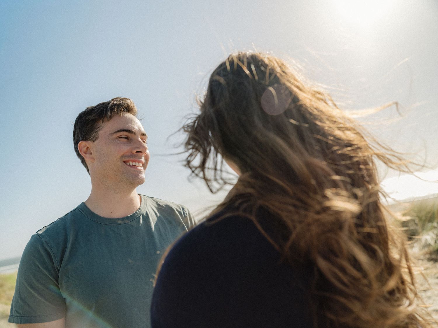 A smiling young man in a teal shirt enjoys a sunny, windy day outdoors with a woman facing away from the camera.