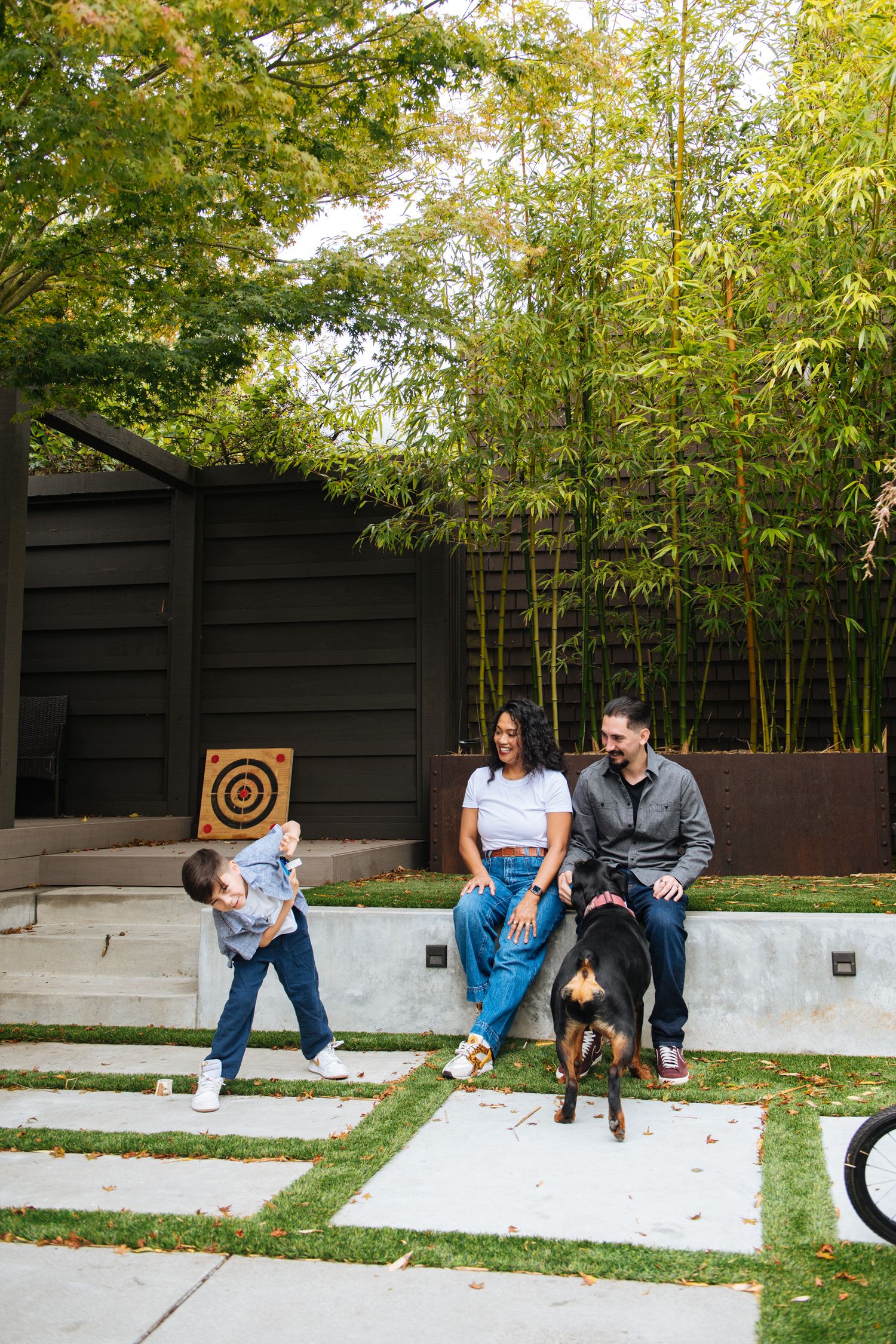 Family with young child and dog relaxing in modern backyard with bamboo, concrete pavers, and lush green turf.
