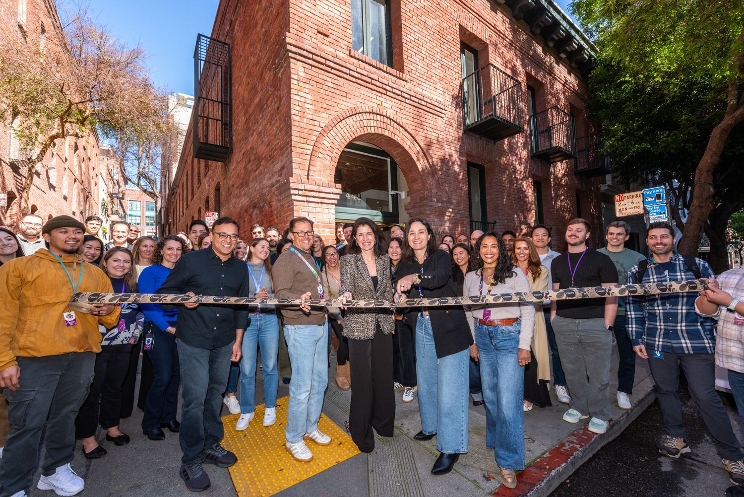 Large group of people celebrating a ribbon cutting ceremony outside a brick building on a sunny day.