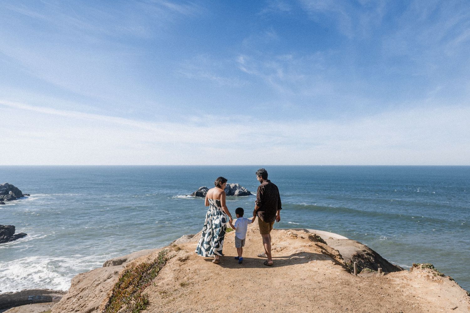 Family of three standing on rocky coastal cliff overlooking the ocean on a sunny day.
