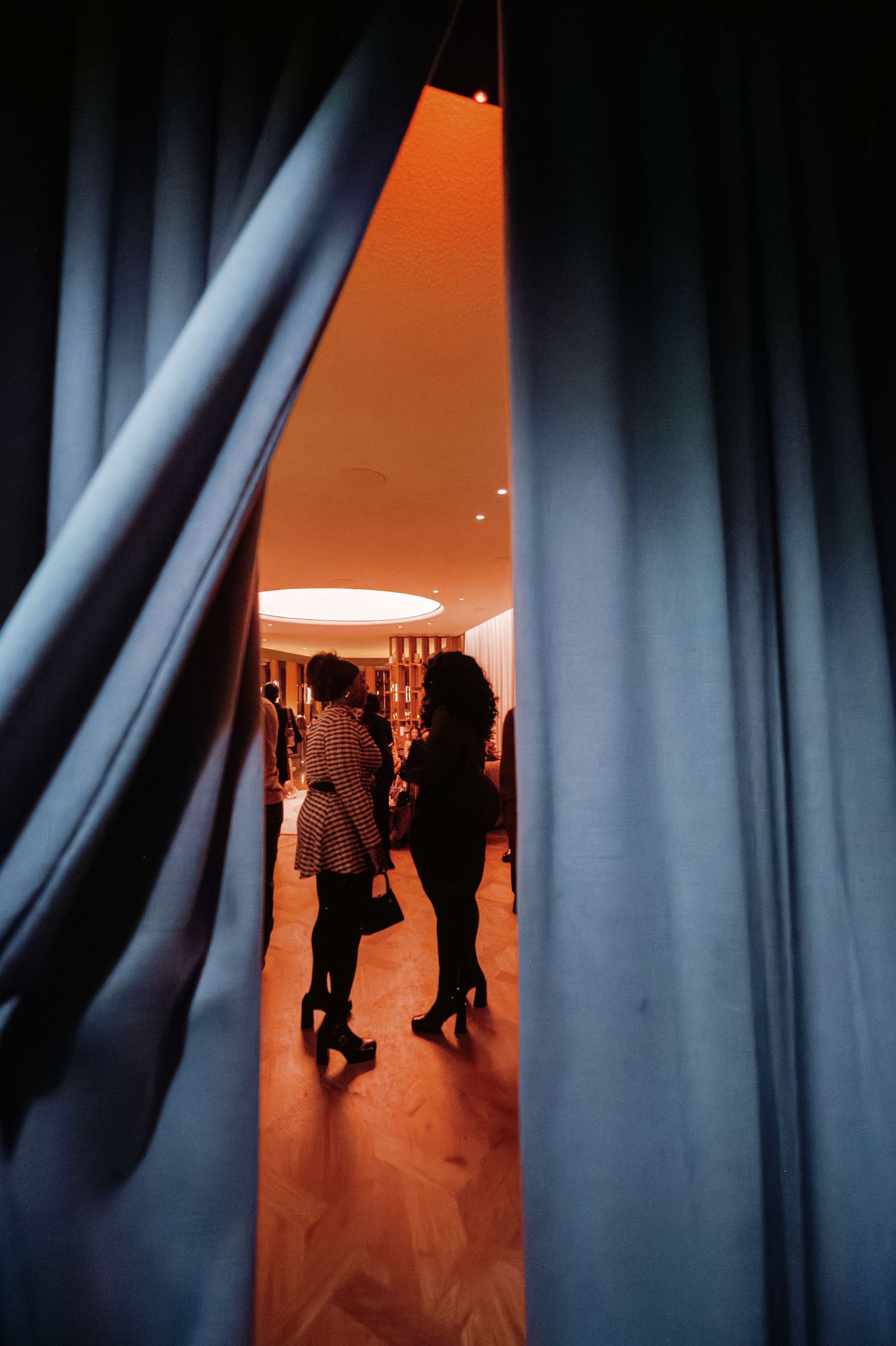 Two women silhouetted in warm light through blue curtains at an elegant indoor social gathering.