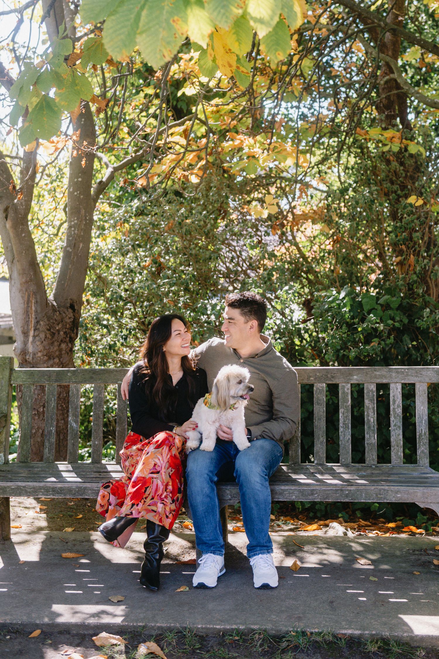 Couple sitting on a wooden bench with a white puppy in an autumn park setting.