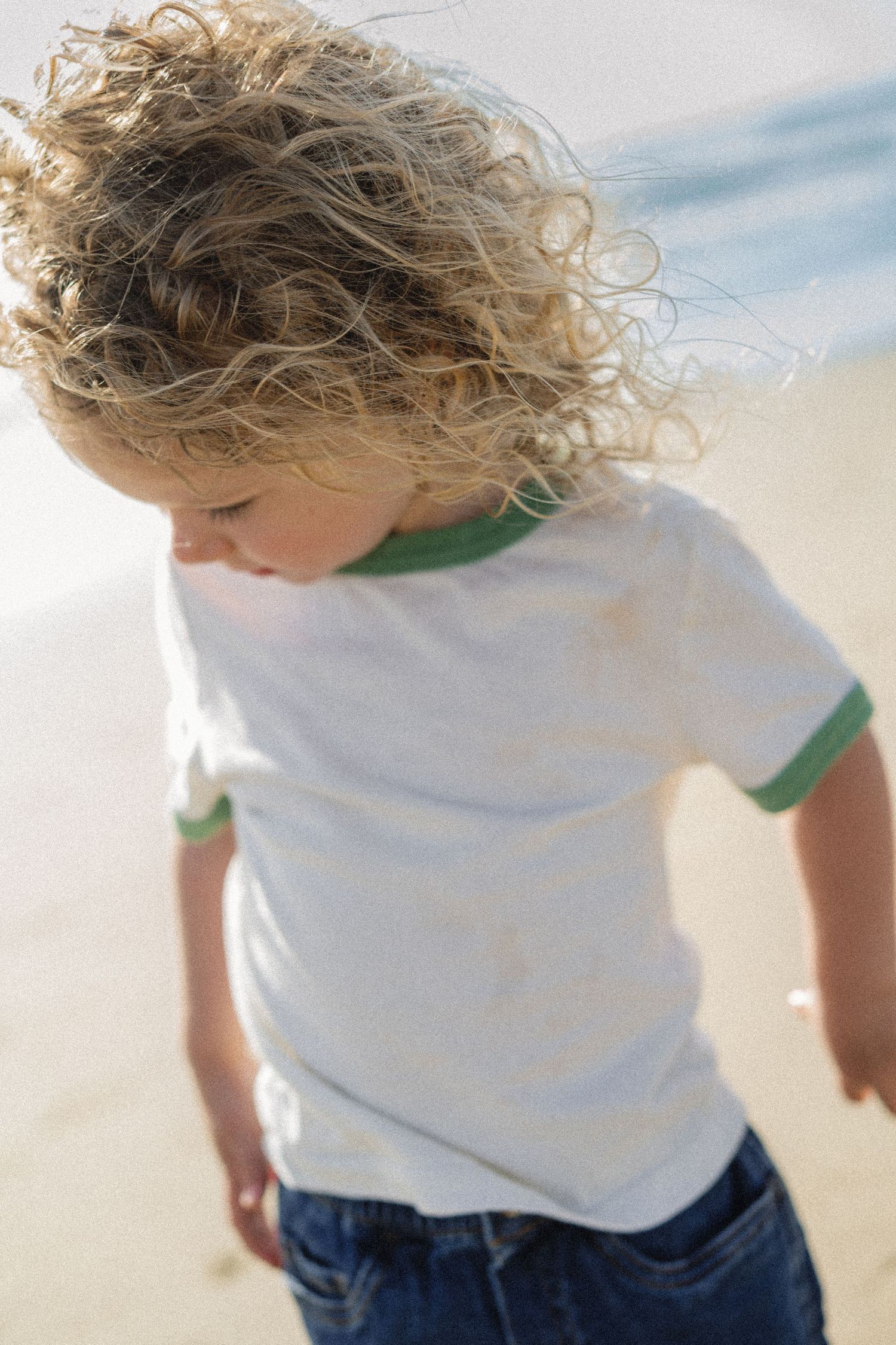 A young child with curly blonde hair wearing a white and green t-shirt and jeans playing on a sunny beach.