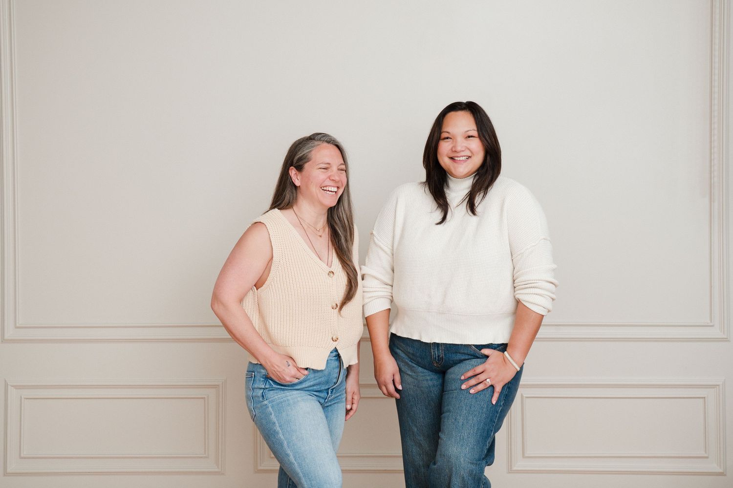 Two women smiling and posing together against a light gray wall, wearing casual jeans and tops.