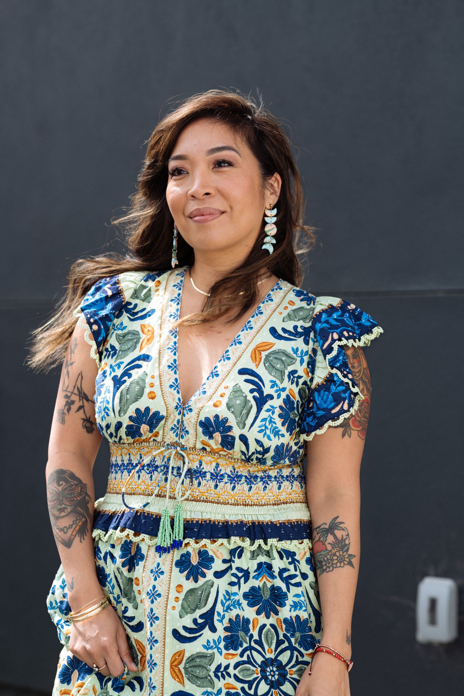 Woman in blue floral dress with tattoos and dangling earrings smiling outdoors.