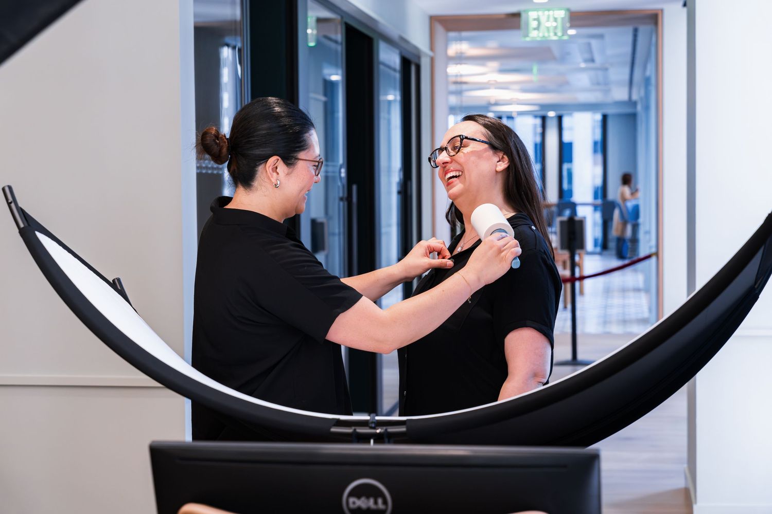 Two women in black outfits laugh together during a professional photo shoot setup with a Dell monitor and reflector.