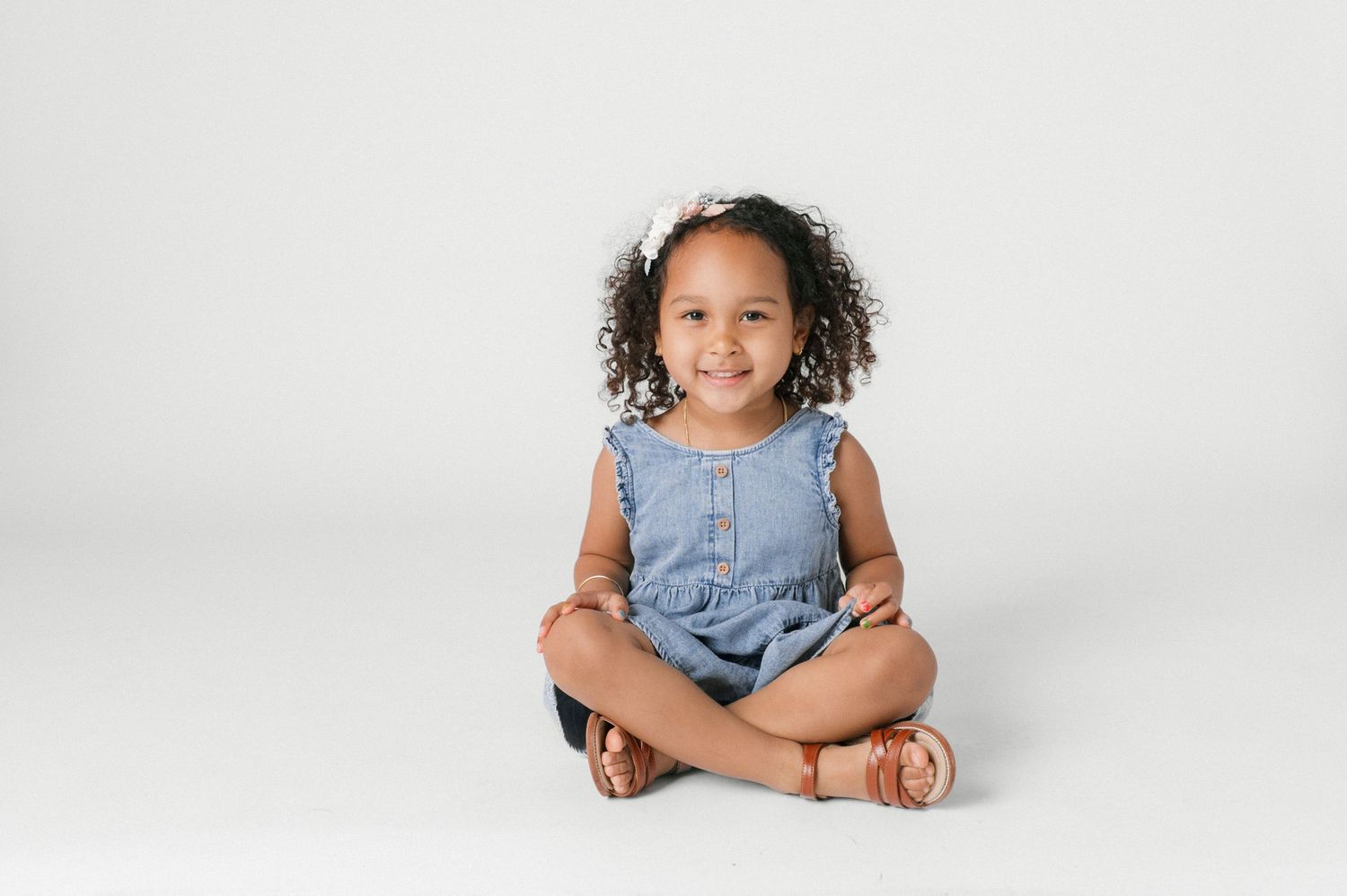 A young girl with curly hair sits cross-legged on a white background, smiling in a blue denim dress.