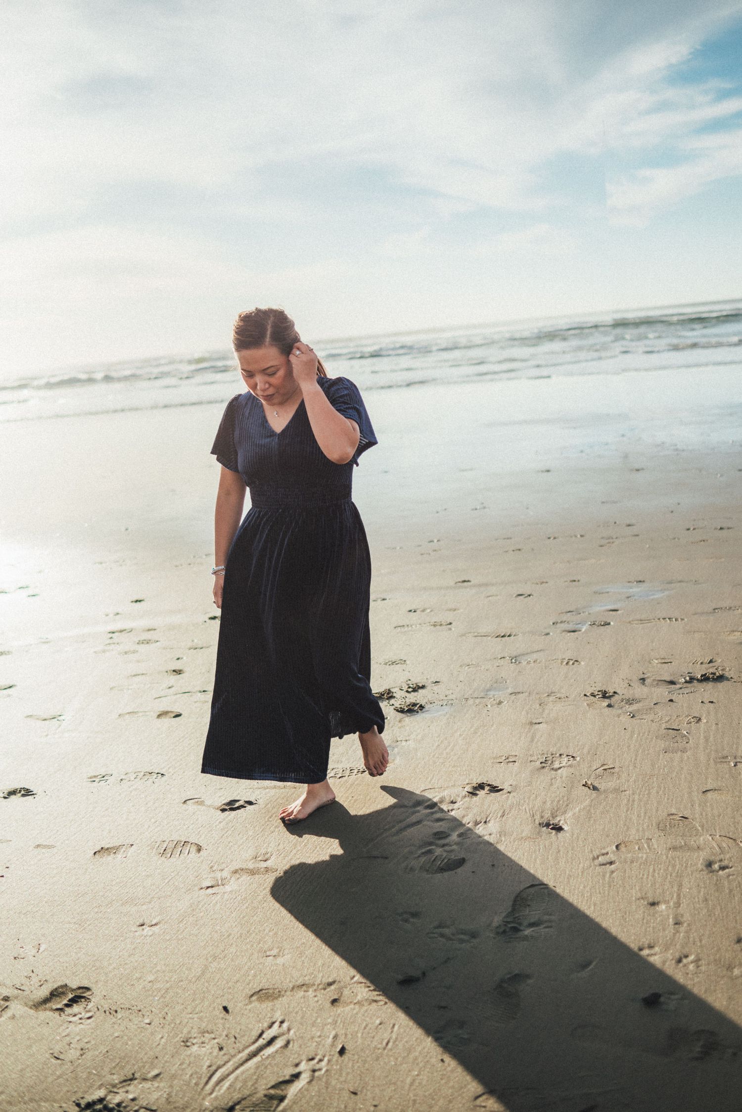 A person in a black dress walks along a sandy beach leaving footprints behind.