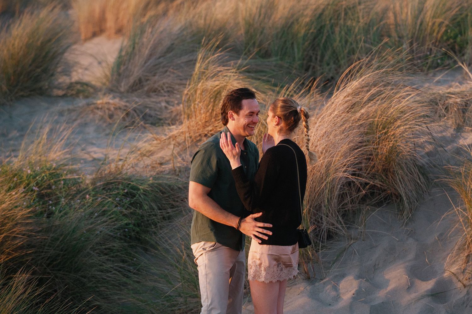 Two people embrace joyfully among coastal sand dunes at dusk.