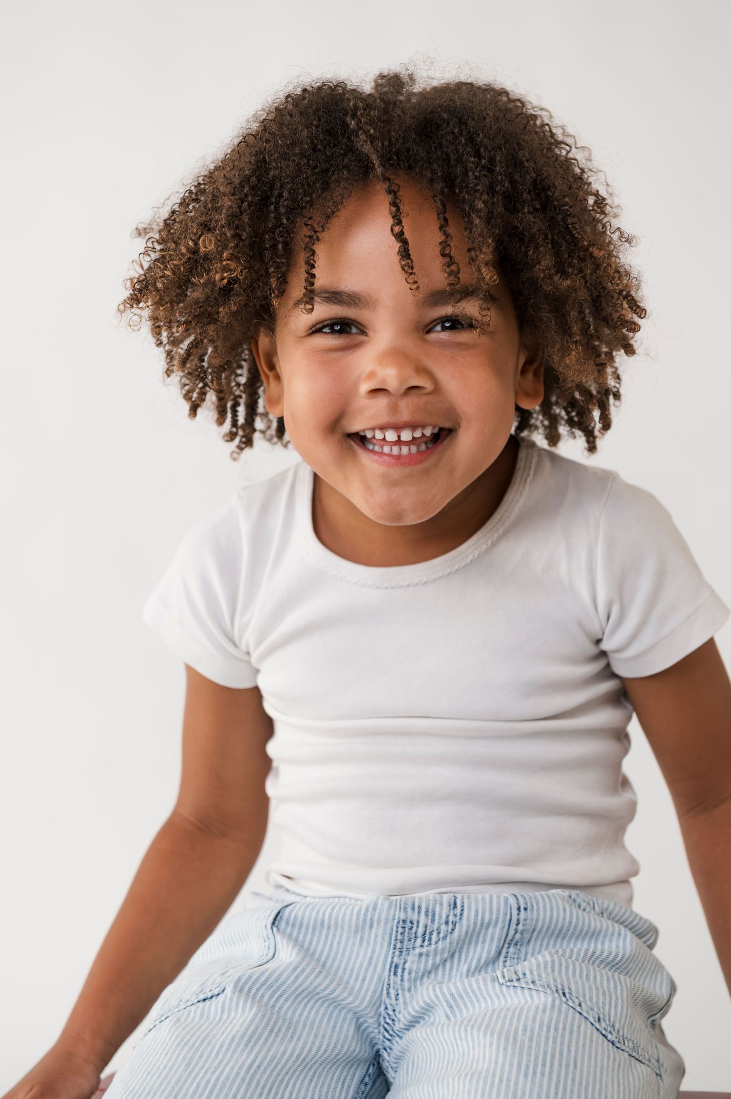 A cheerful young child with curly hair smiles brightly, wearing a white tee and light jeans against a white background.