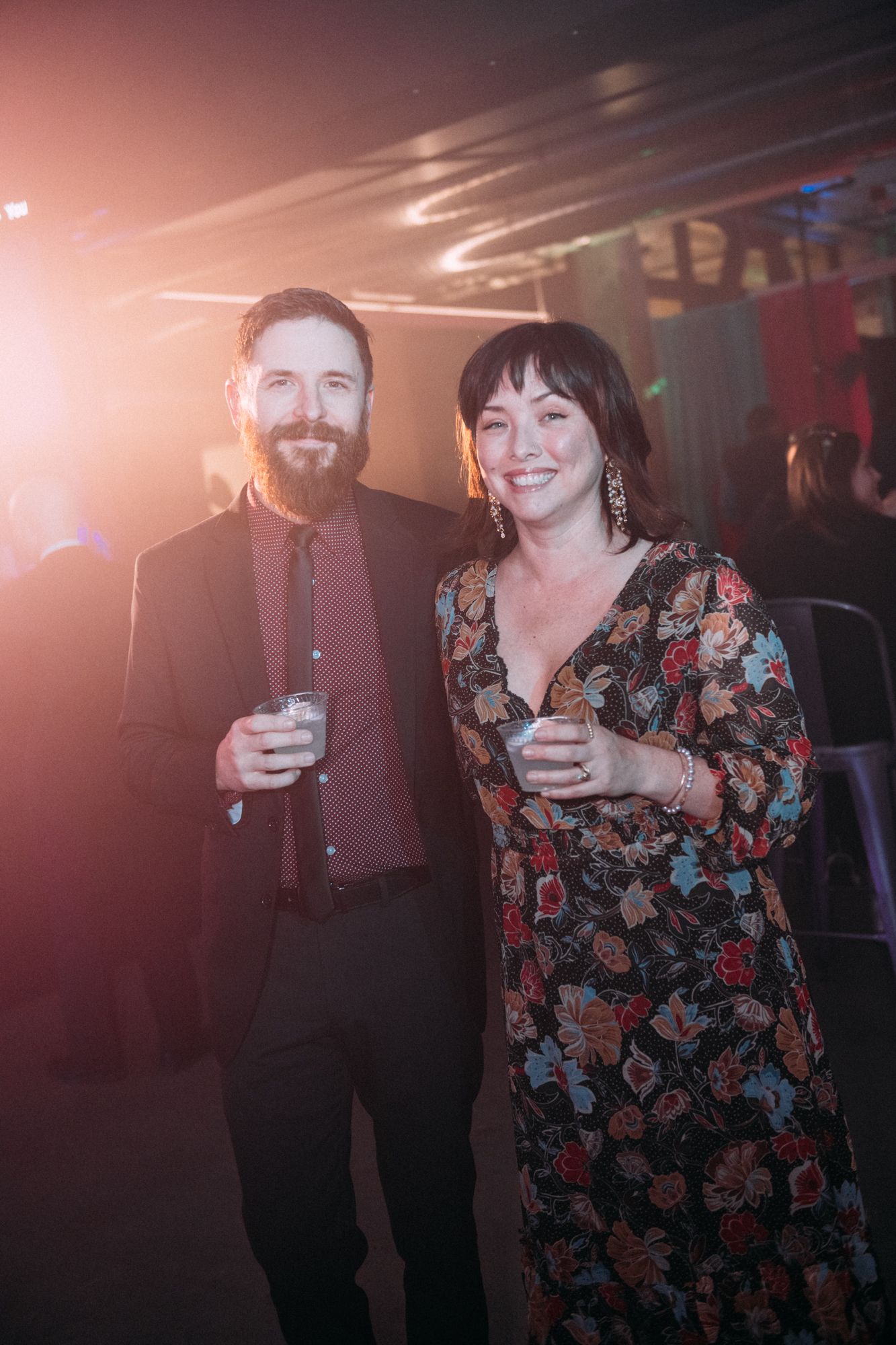 A couple stands together at a party, with one person wearing a floral dress and another in dark clothing in dim lighting.