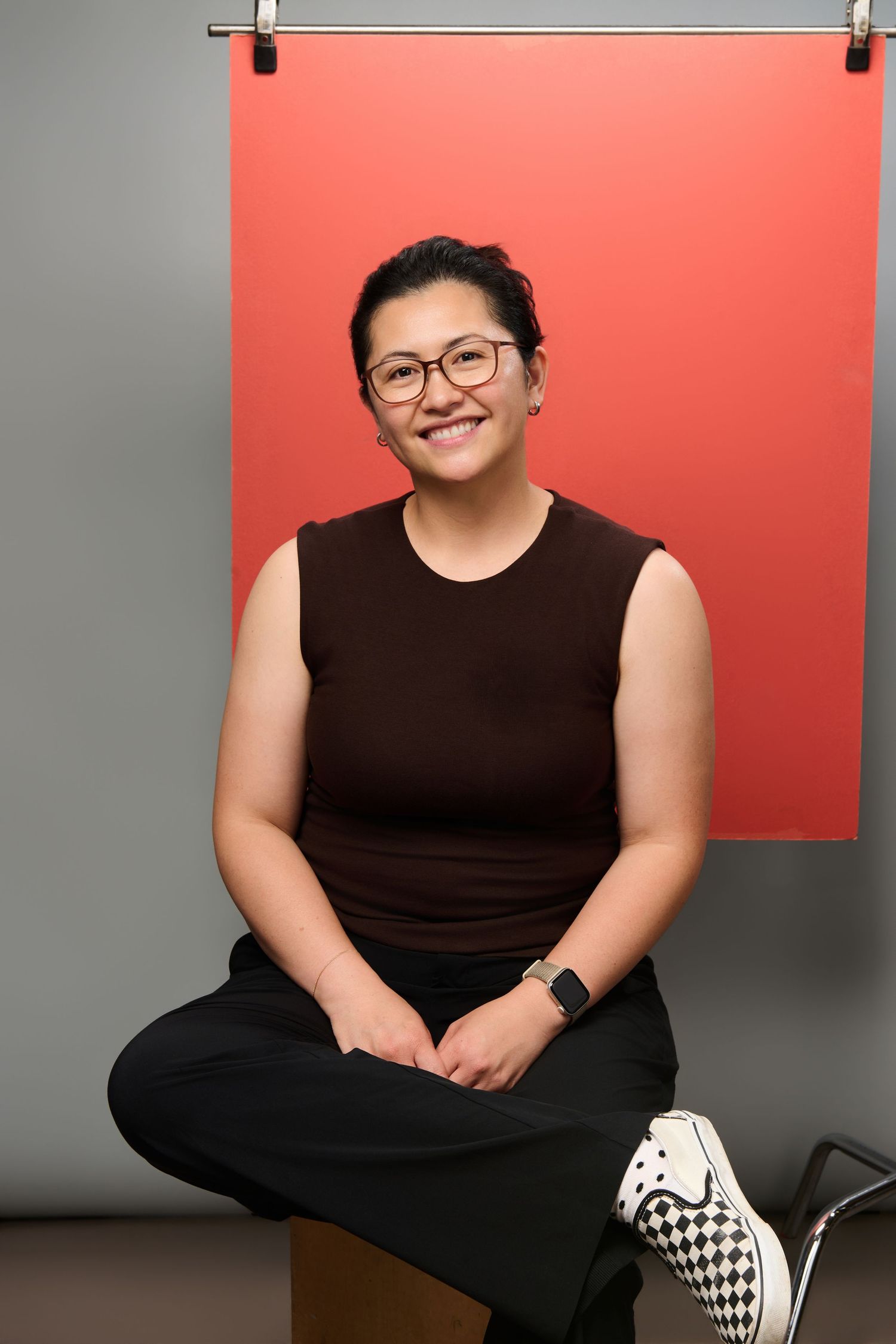 Smiling person in brown sleeveless top and black pants sits cross-legged before a red backdrop.