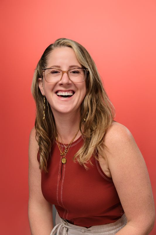 Smiling woman with glasses and layered necklaces wearing a red top against a coral background.