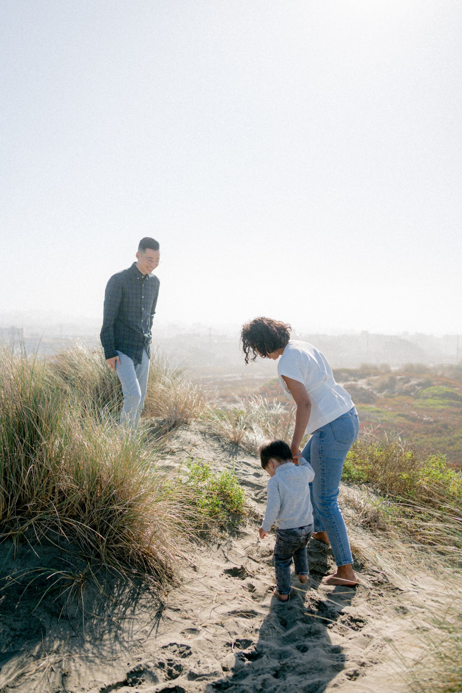Family of three enjoying a sunny day on sandy beach dunes, with parents guiding their toddler along the path.