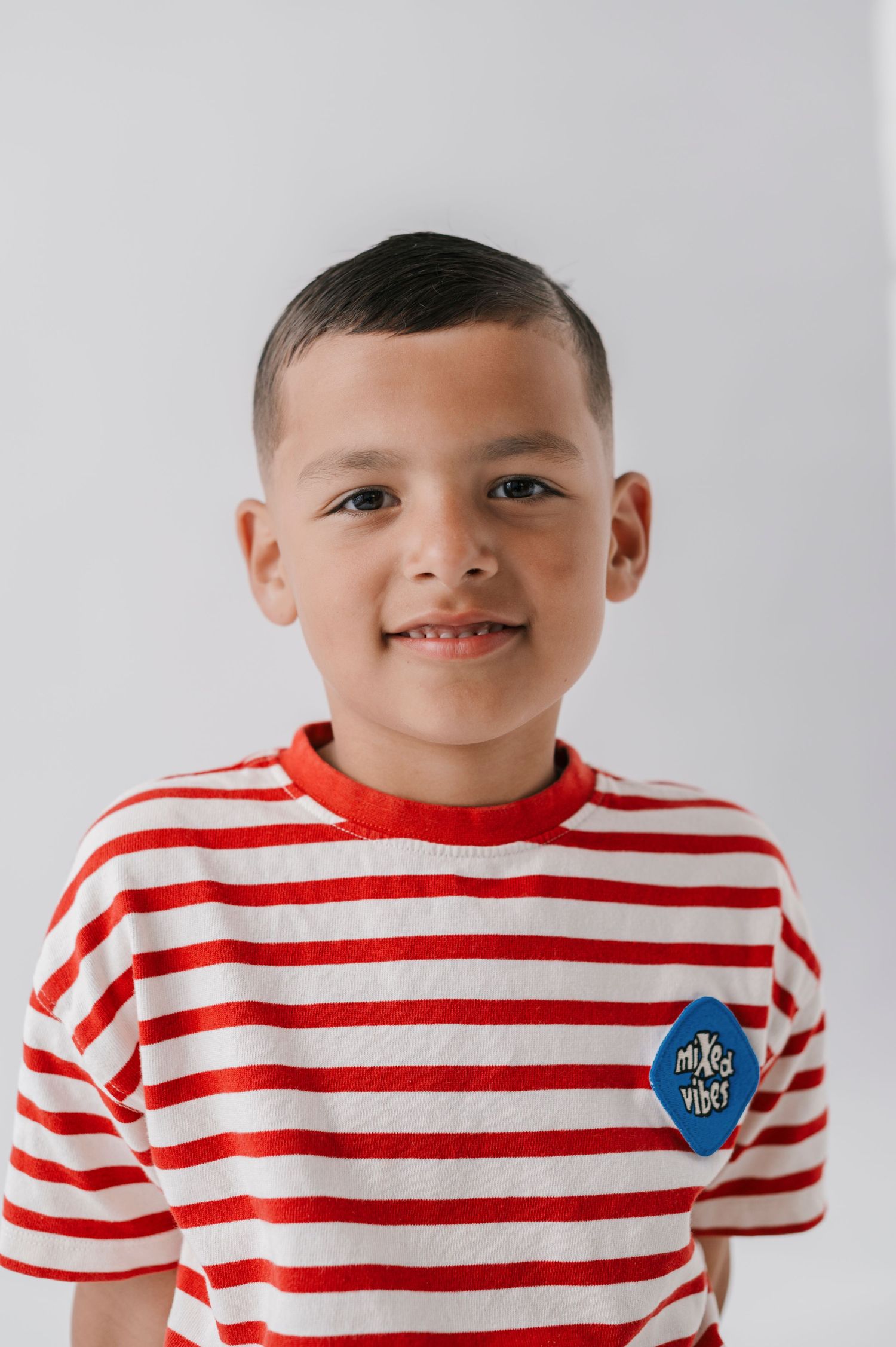 Smiling young boy wearing a red and white striped t-shirt with a blue badge, photographed against a white background.