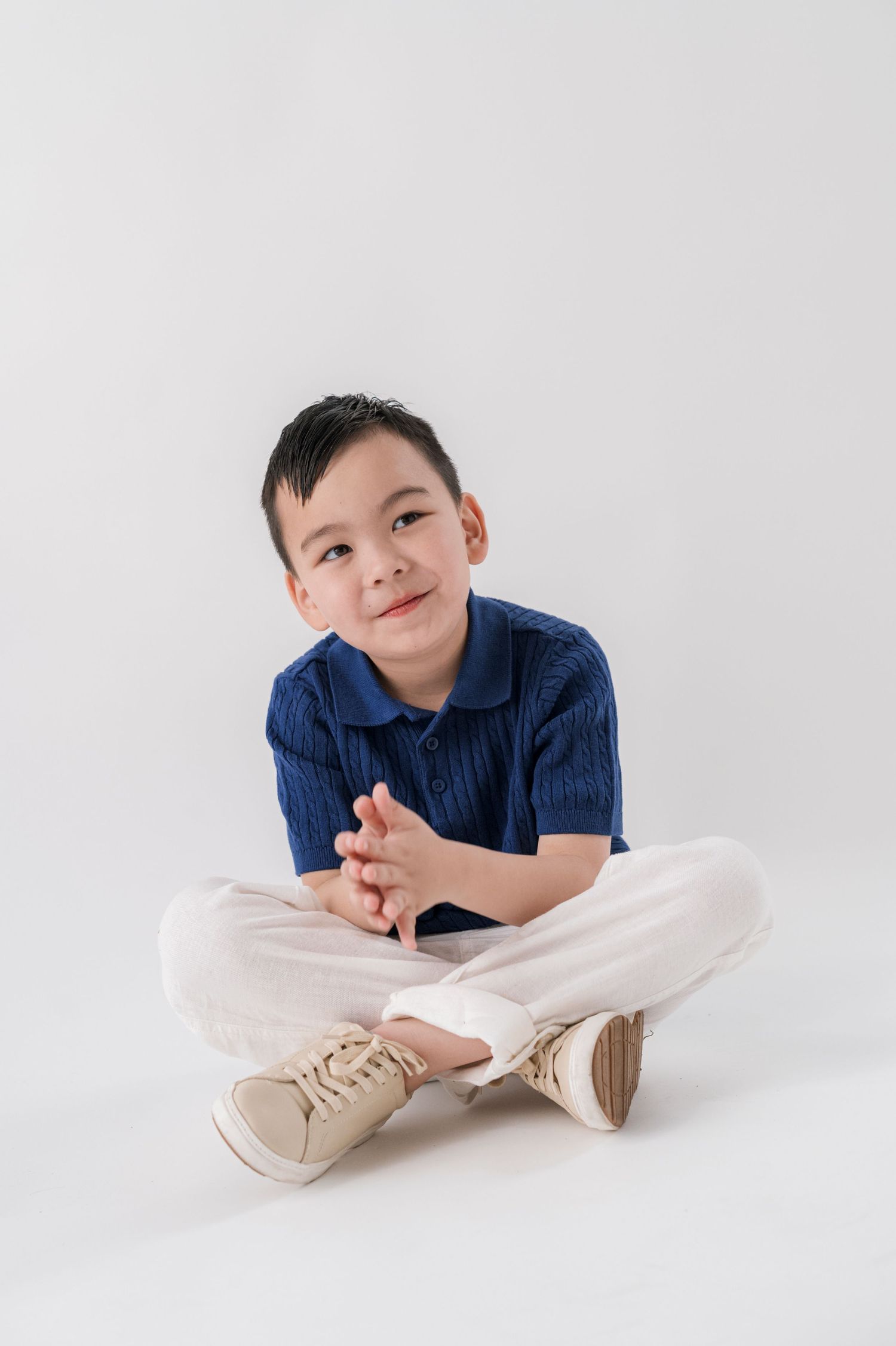Young Asian boy sitting cross-legged on white background, wearing blue polo shirt and white pants, smiling thoughtfully.