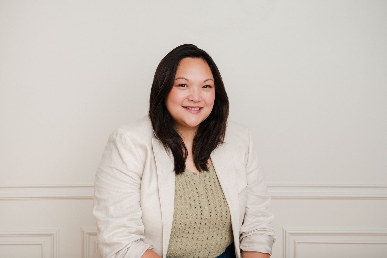 Smiling woman in white blazer and green top poses confidently against a light gray background.