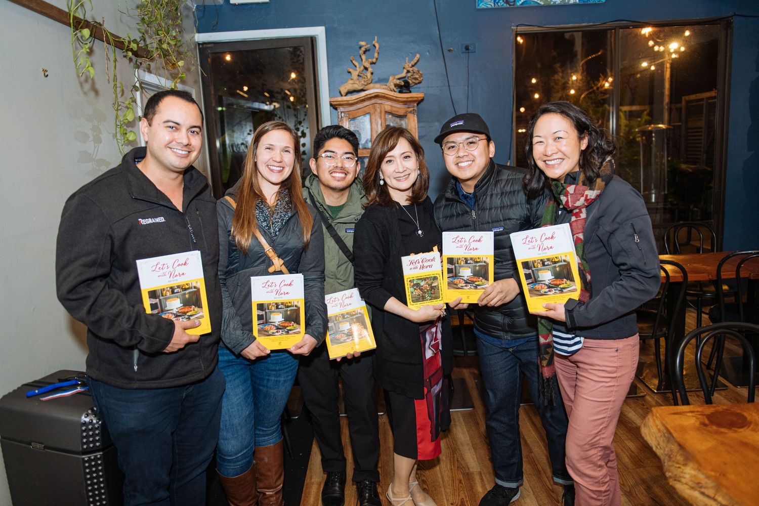 A group of people stand together at an event holding books and smiling for a photo in a casual indoor setting.