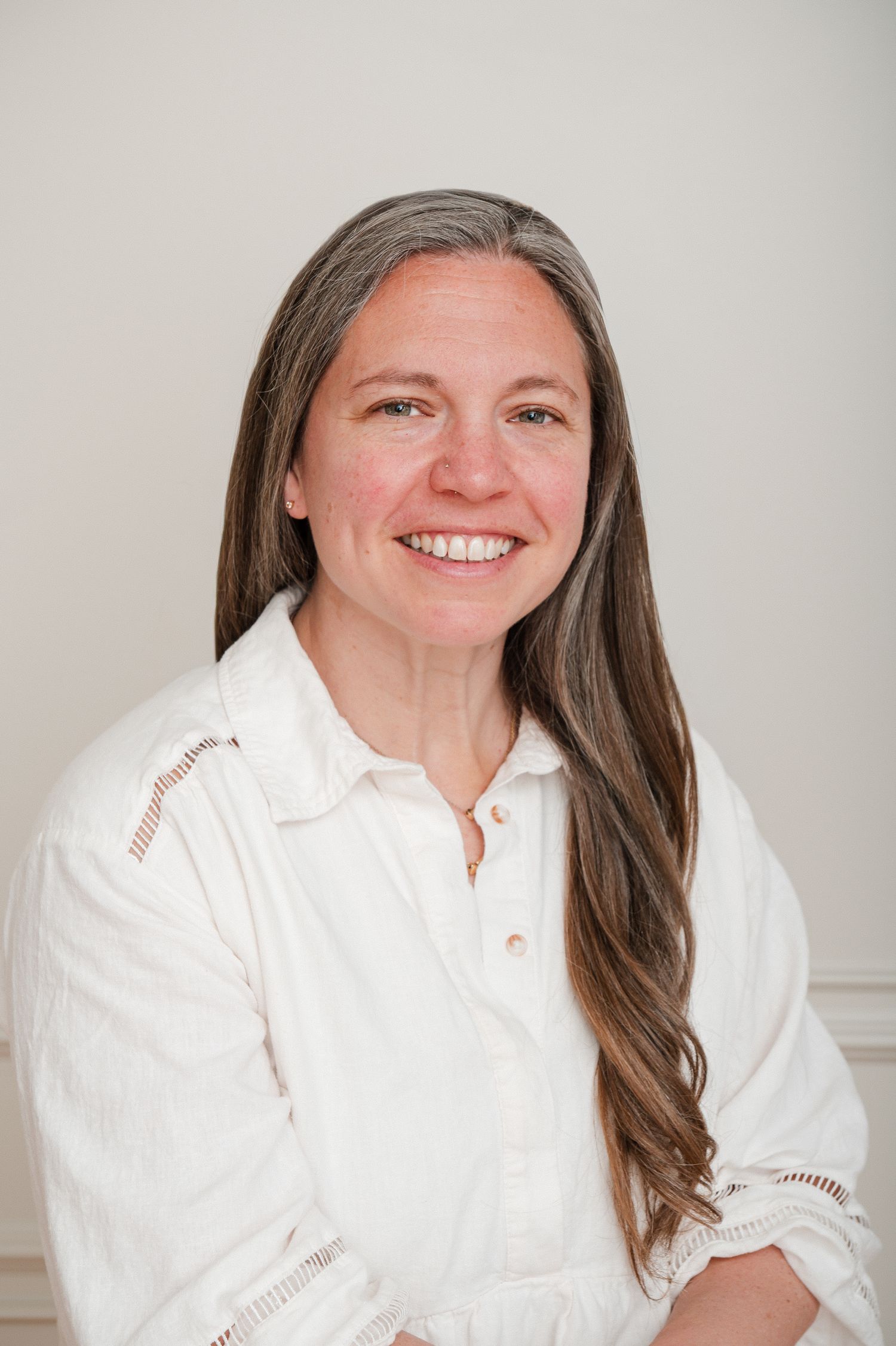 Smiling woman with long gray-streaked hair wearing a white embroidered blouse, posed against a light background.
