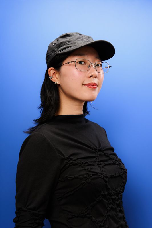 Young woman in black top, gray cap, and glasses posing against a blue background.