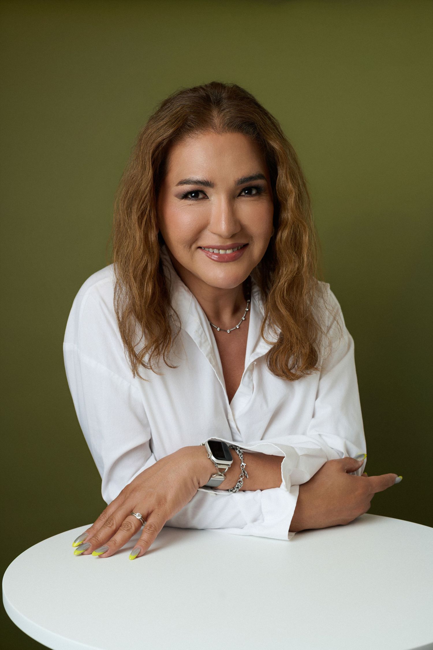 A smiling woman in a white shirt leans on a white table, wearing a smartwatch and ring, against an olive background.