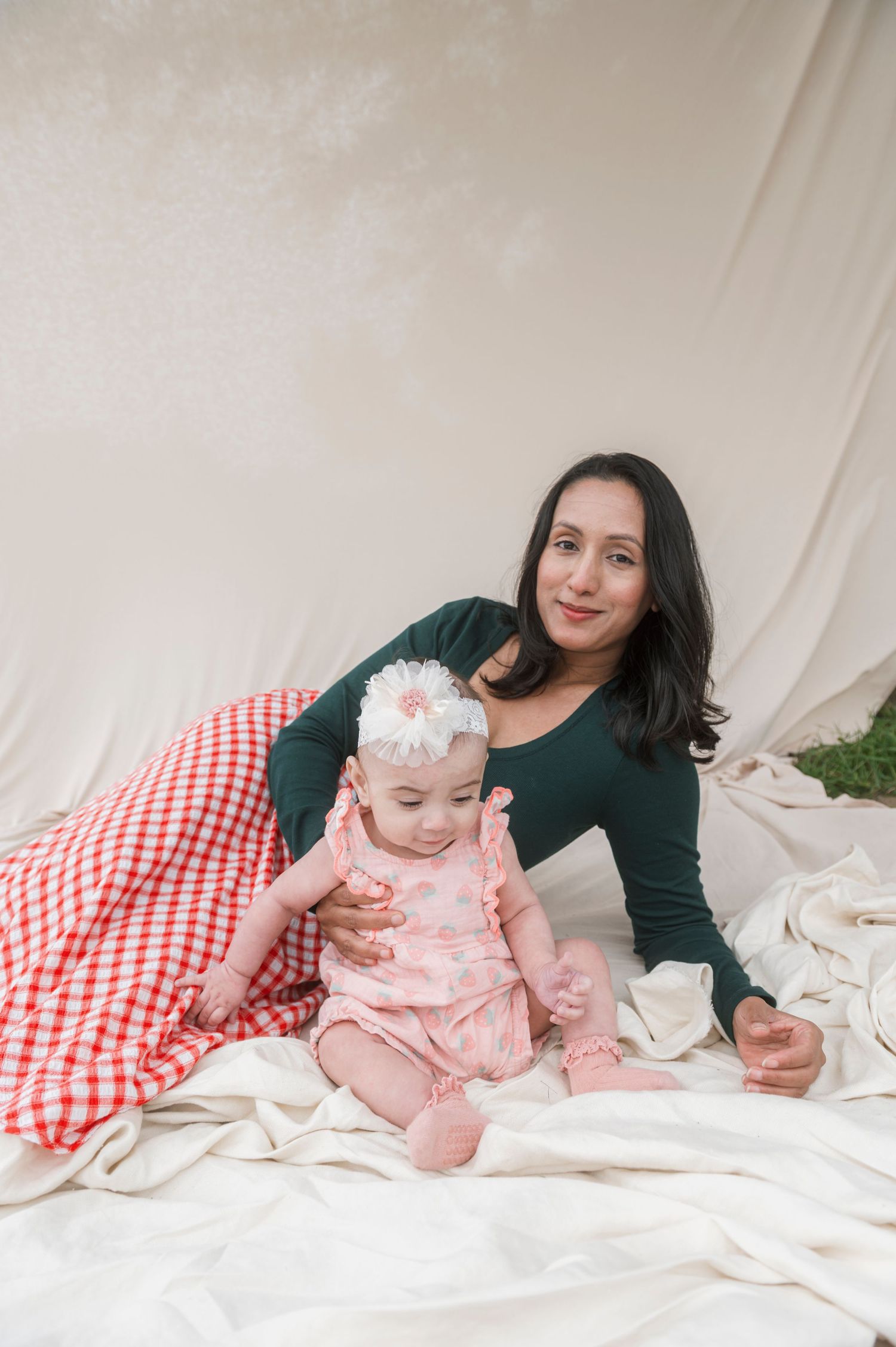 A smiling woman in green sits with a baby girl in pink floral outfit and white headband on white blankets.