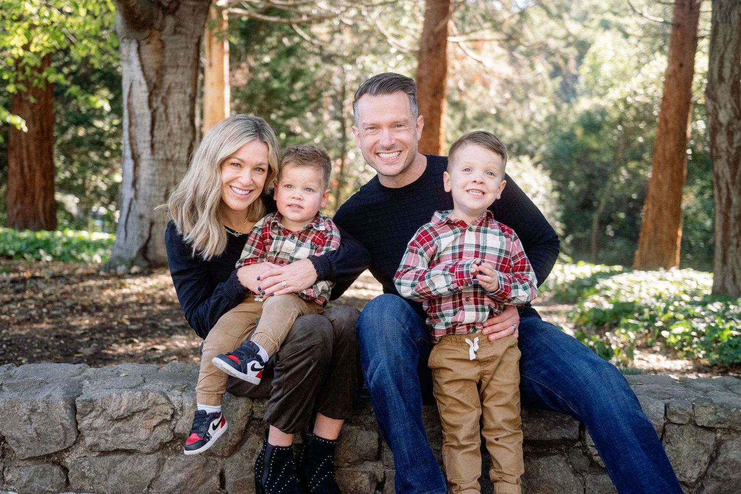 Family wearing matching plaid outfits sitting together on stone wall in wooded setting.