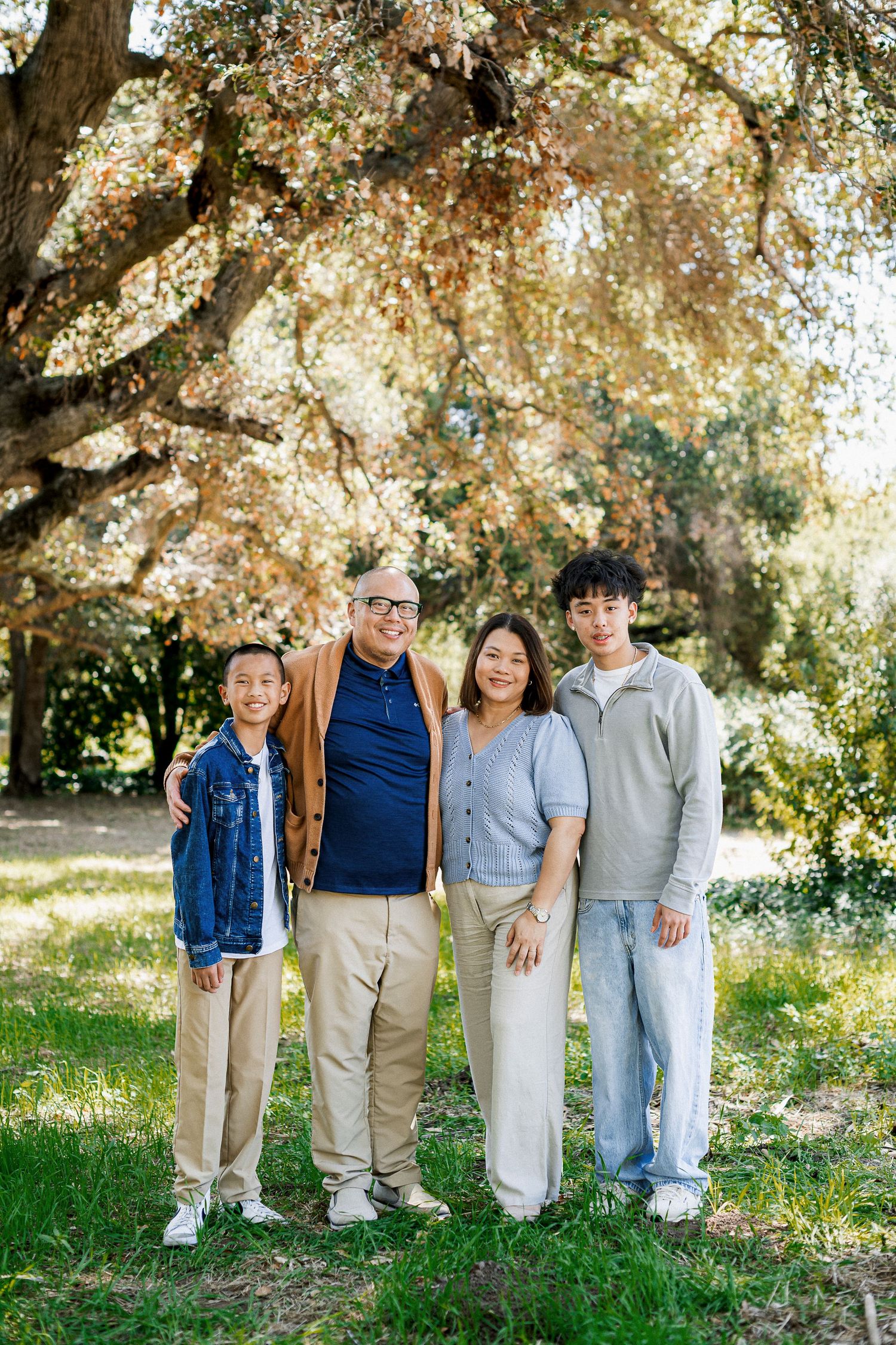 Asian family of four smiling together outdoors under a large tree in a sunny park setting.