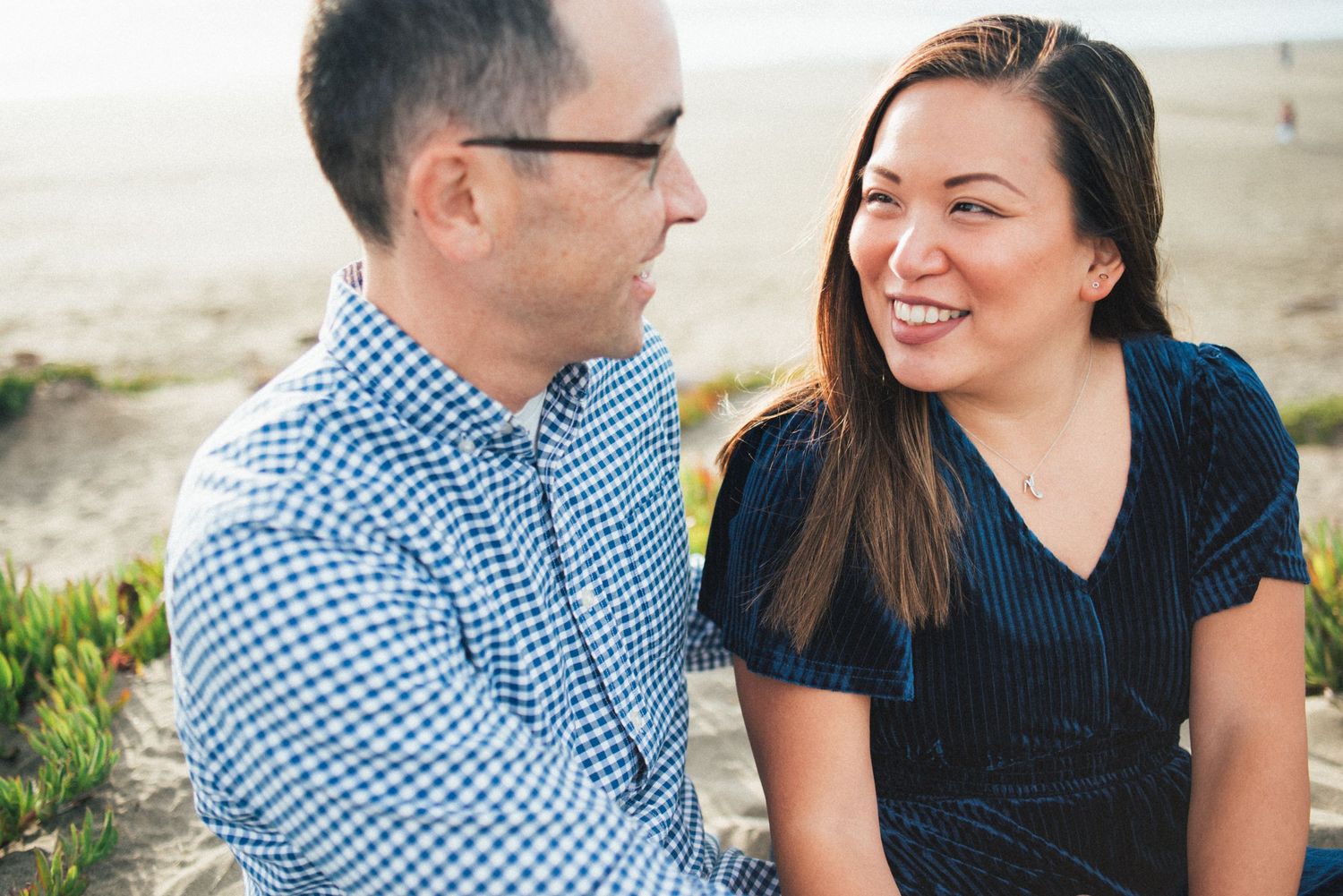 A couple shares a joyful moment together on a sunny beach with mountains in the background.