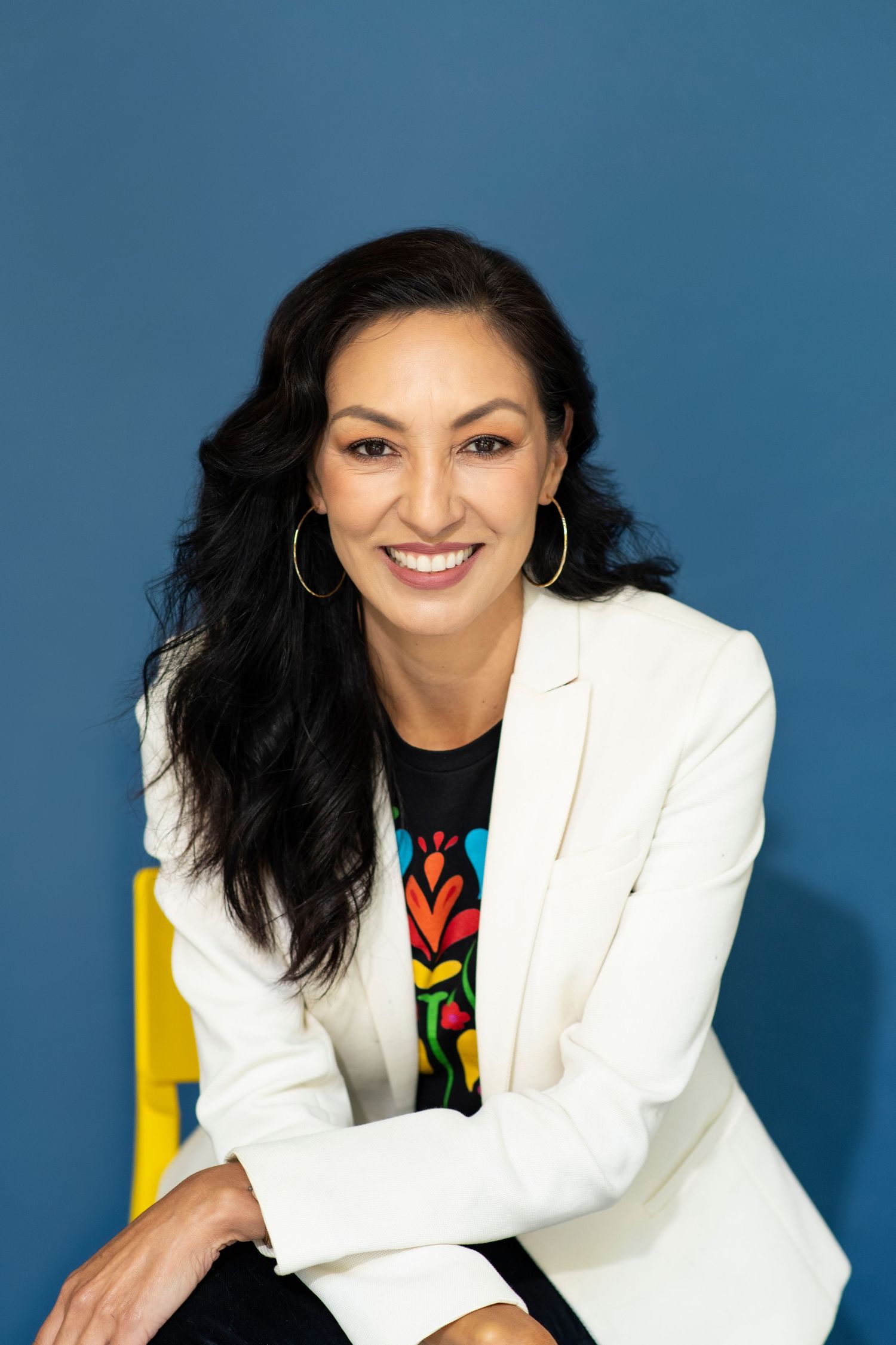 Smiling woman with dark hair in white blazer and colorful top, seated against blue background.