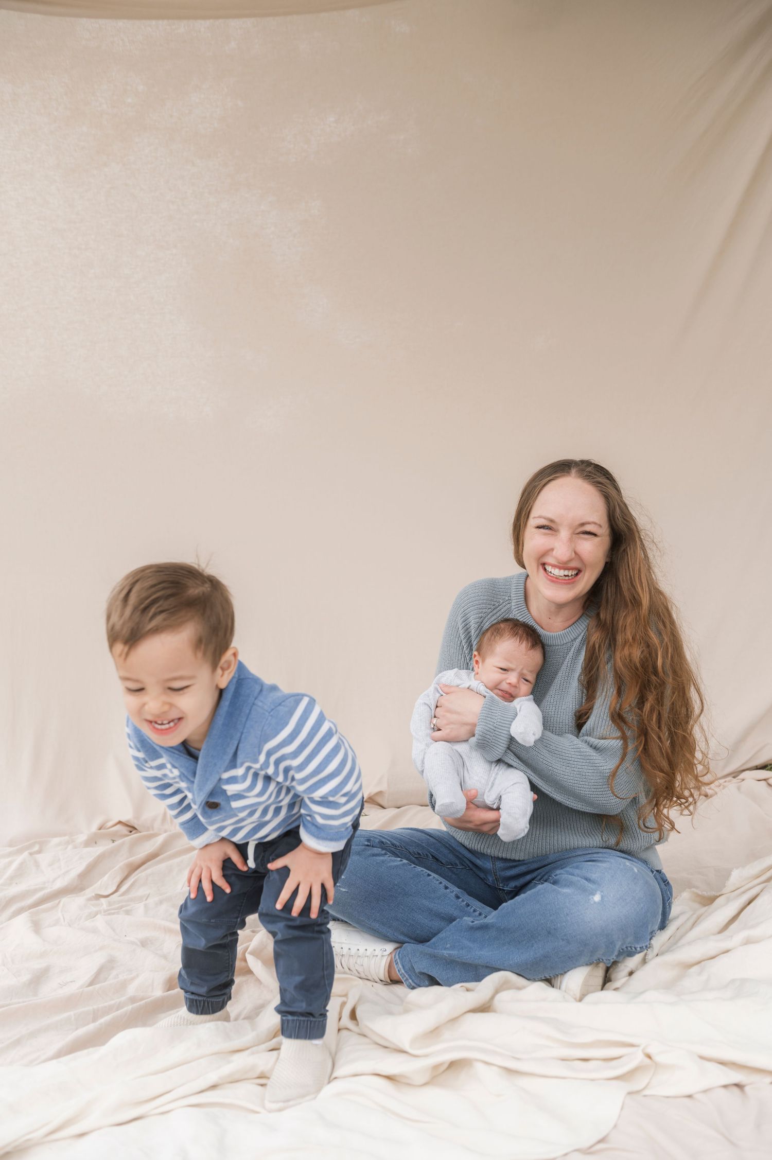 A smiling mother with long hair holds a newborn while her toddler boy plays beside her on a white blanket.