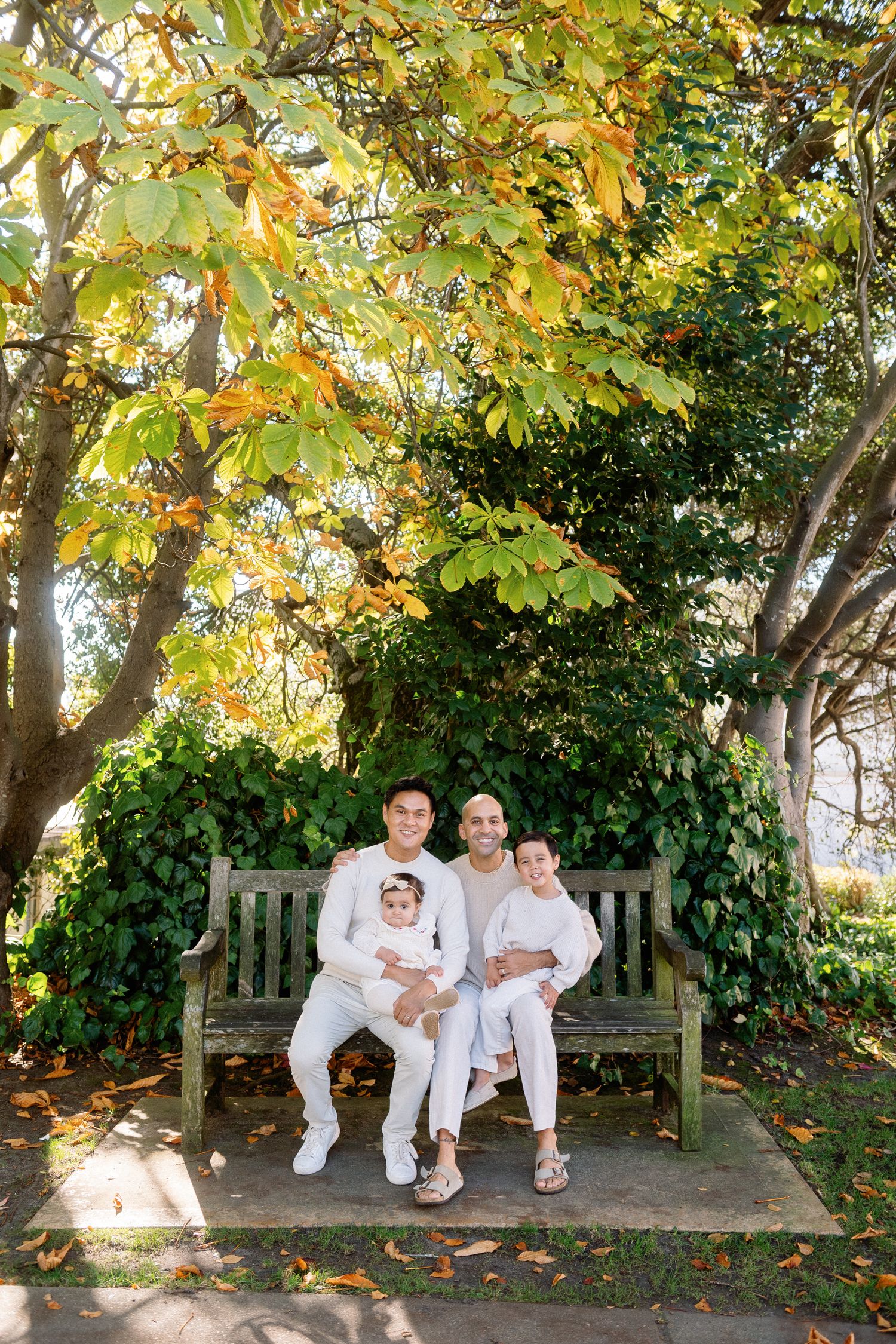 Family dressed in white relaxing on a wooden bench under fall foliage.