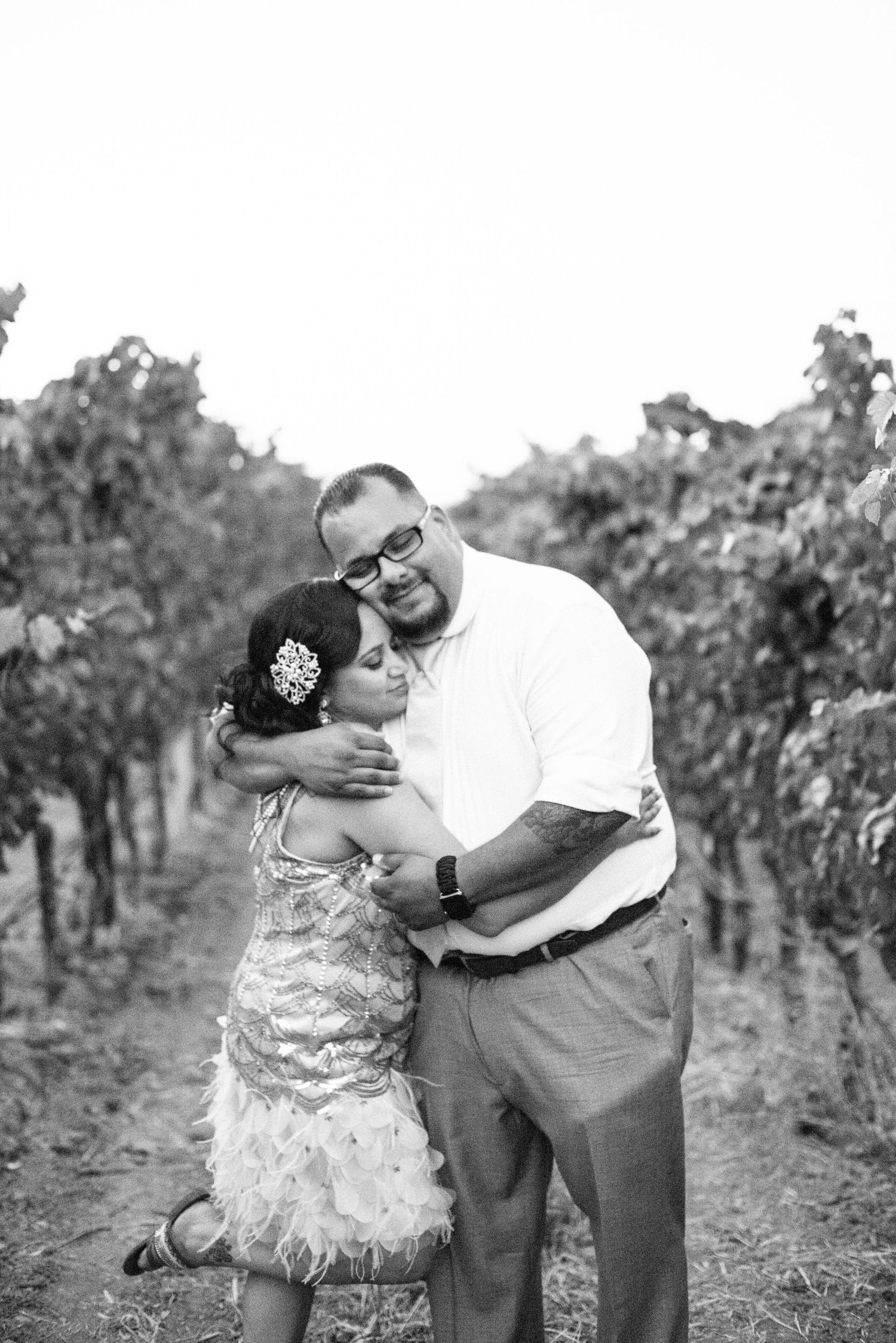 A couple embraces lovingly in a vineyard setting during what appears to be an engagement or wedding photoshoot.