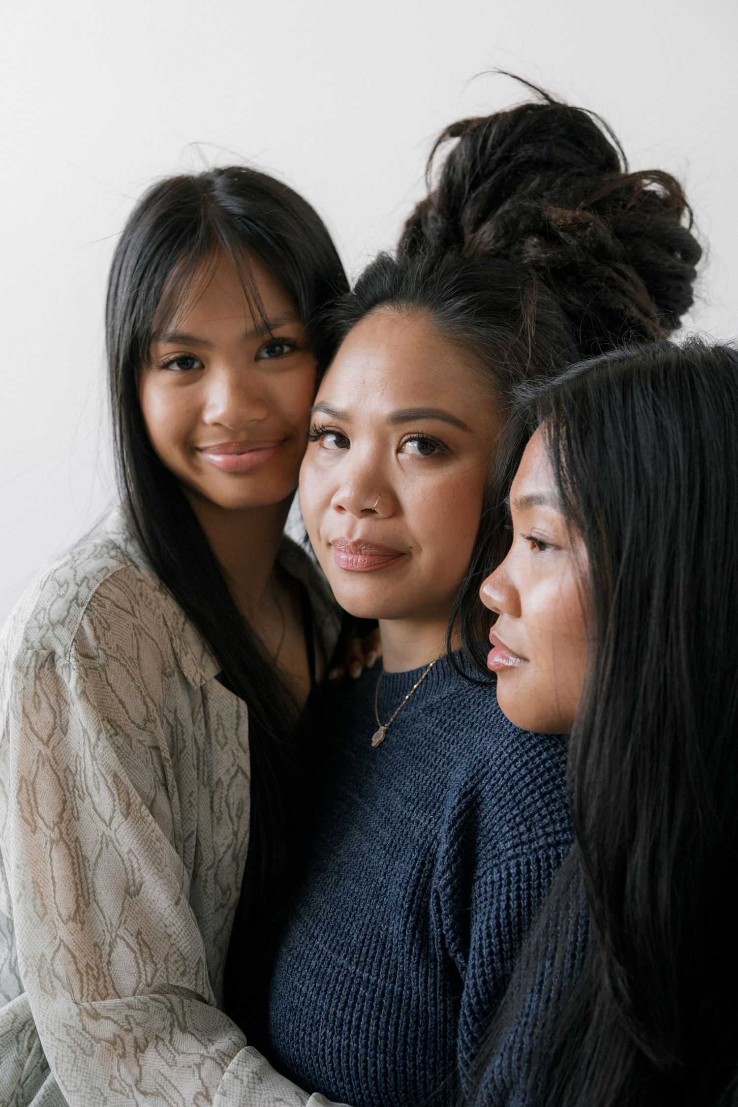 Three young women with dark hair pose together closely, smiling softly against a white background.