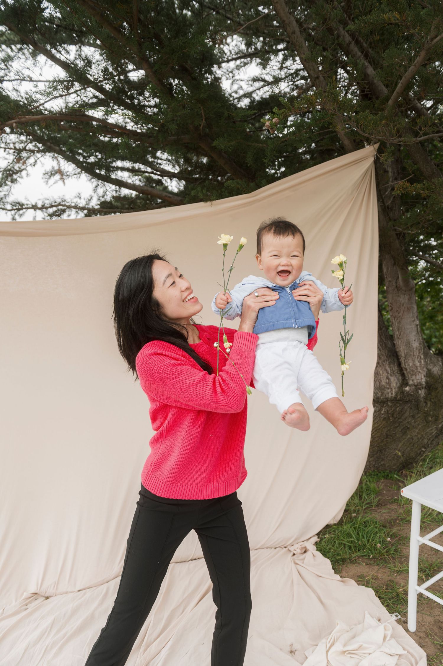 A woman in a red sweater joyfully lifts a laughing baby holding flowers outdoors against a beige fabric backdrop.