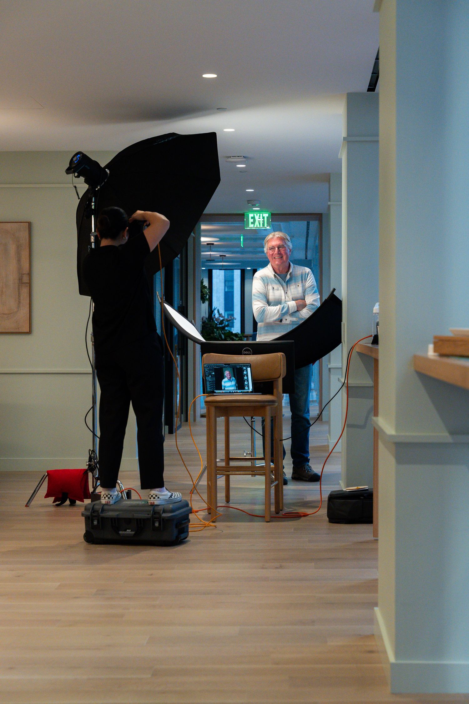 Photographer setting up studio lights to photograph a man posing on a stool in an indoor hallway setting.