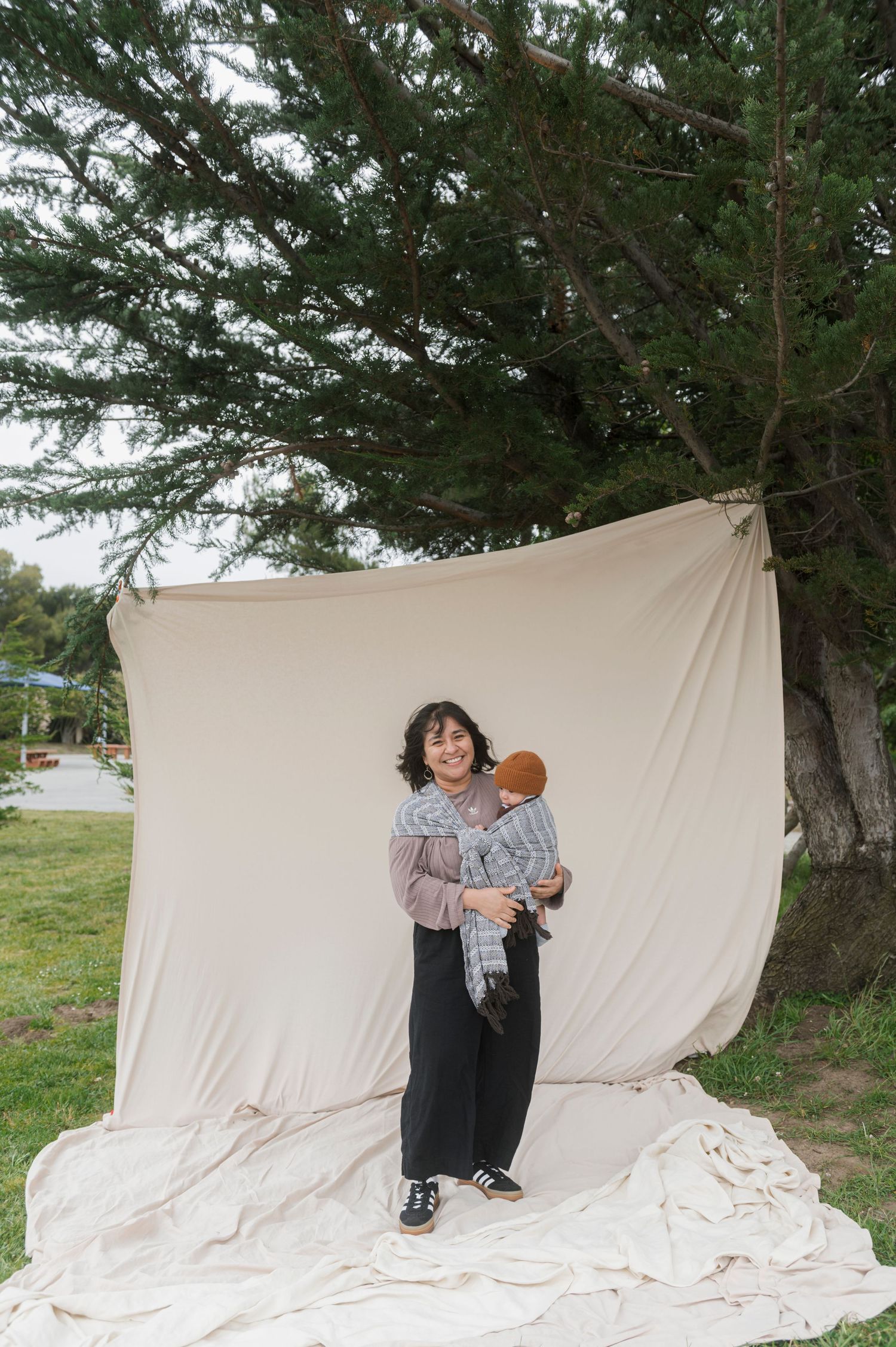 A woman smiles while holding a baby outdoors in front of a white fabric backdrop hung near a large tree.
