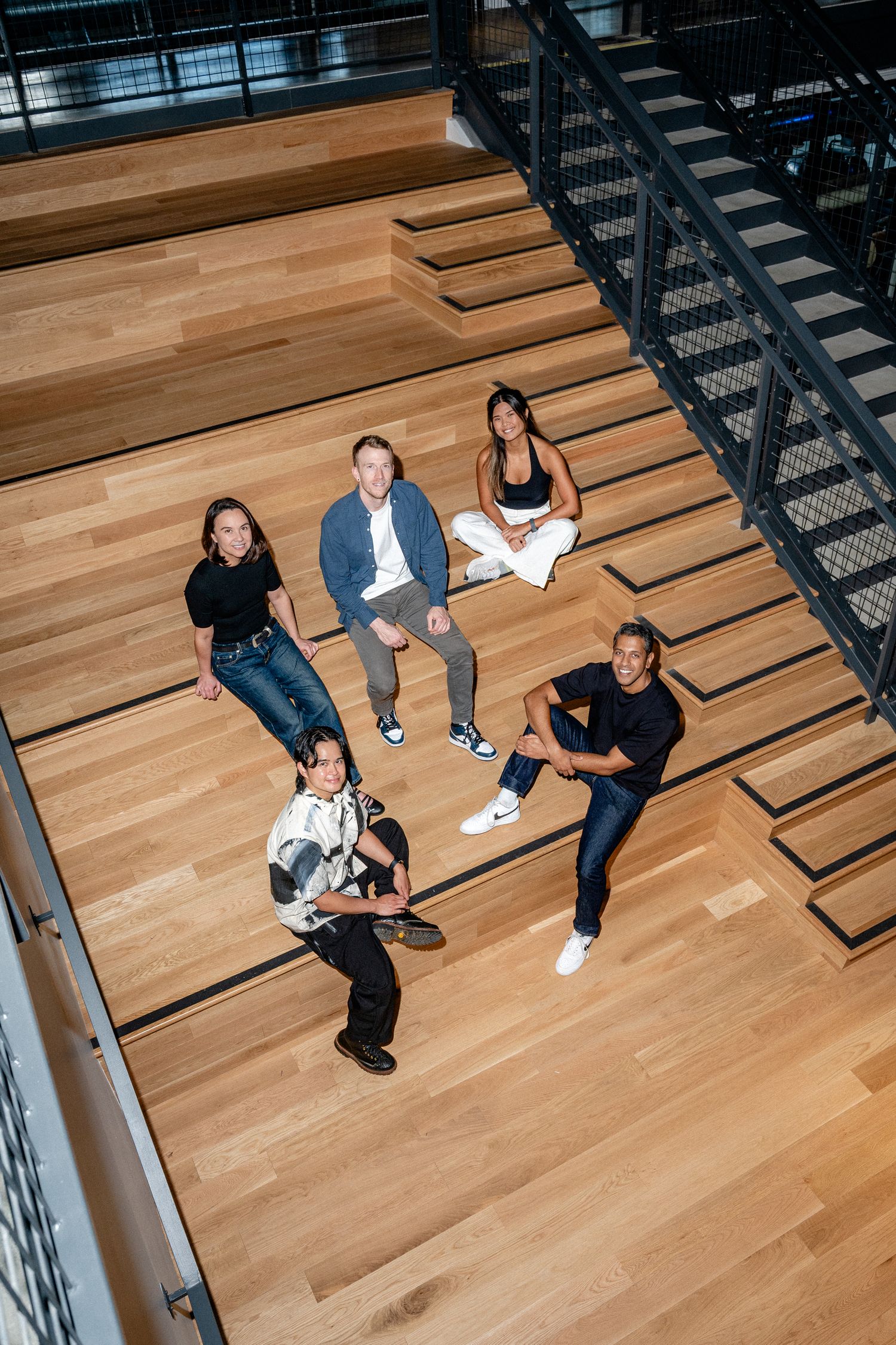 Five diverse professionals posed on modern wooden stadium steps in a contemporary office building, viewed from above.